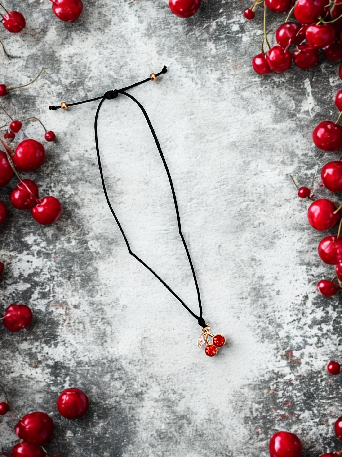 Black bracelet with red cherry charm on a grey surface surrounded by red berries