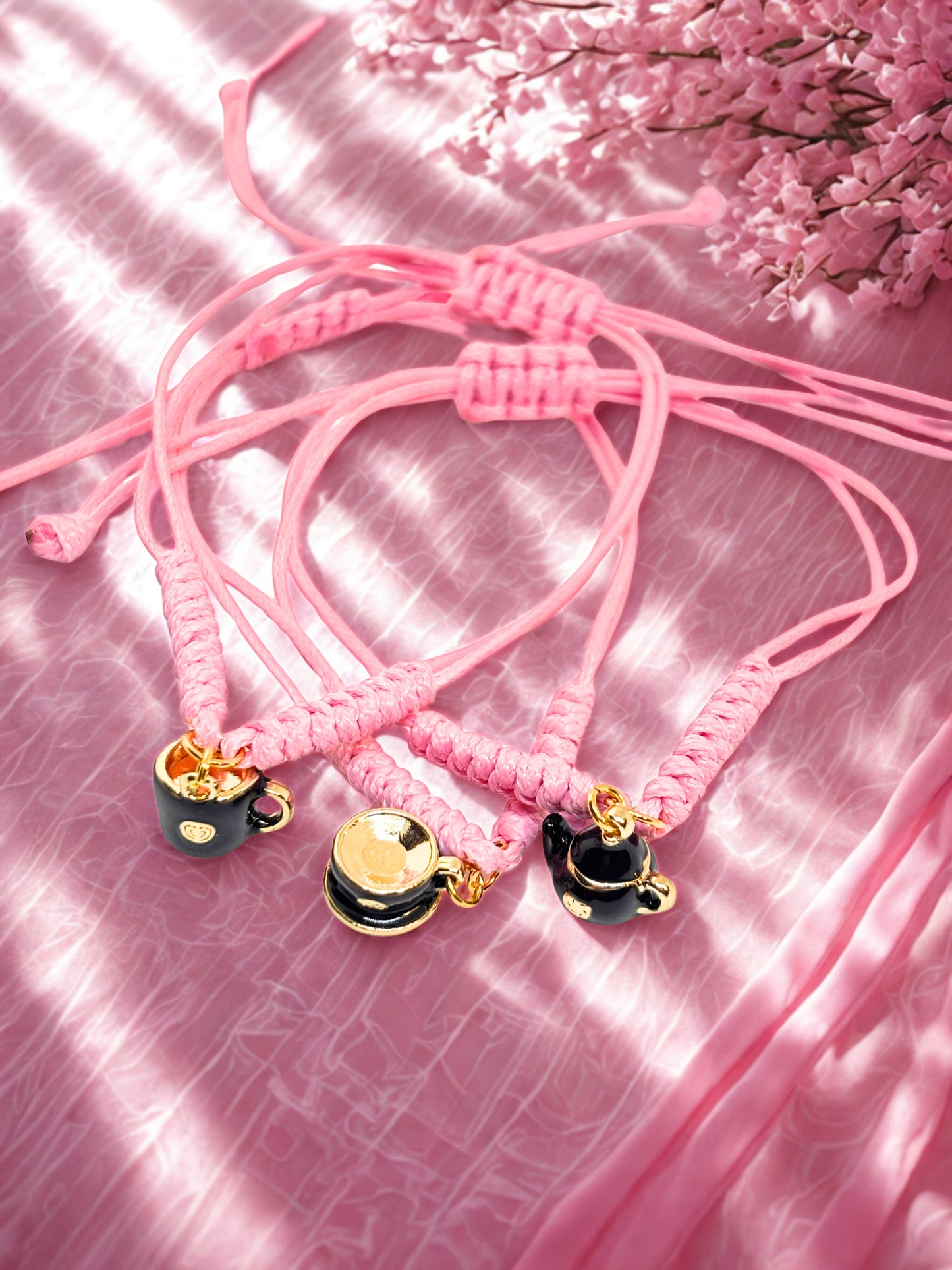 Pink braided bracelets with black and gold teapot and teacup charms on a pink textured background