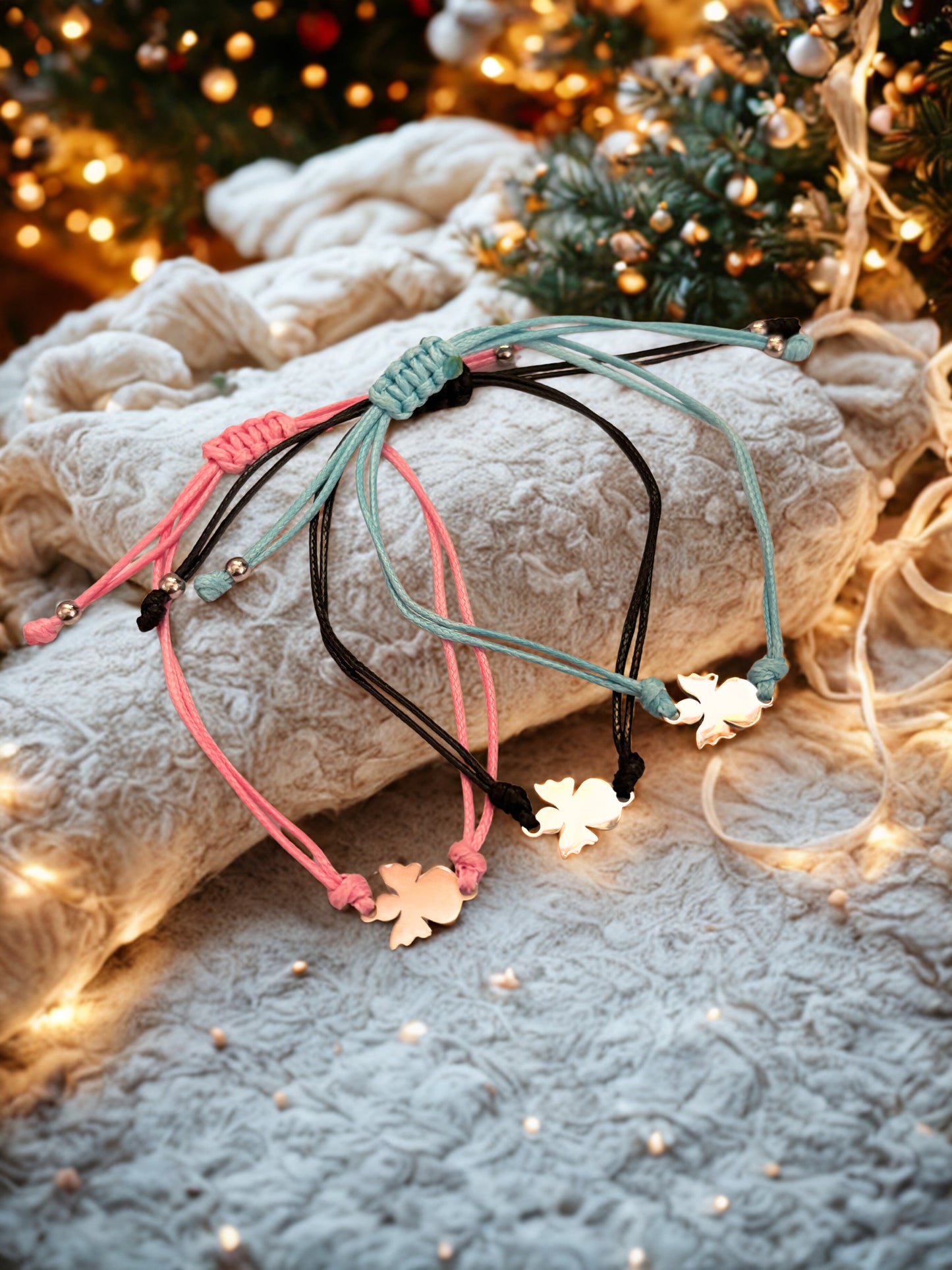 Three braided bracelets with angel charms on a textured surface with Christmas lights and decorations.