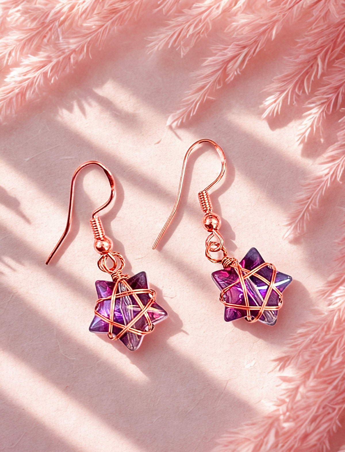 A pair of handmade wire-wrapped dangle earrings with purple faceted star-shaped charms and rose gold hooks, displayed on a textured pink surface with soft dried grass and dappled sunlight.