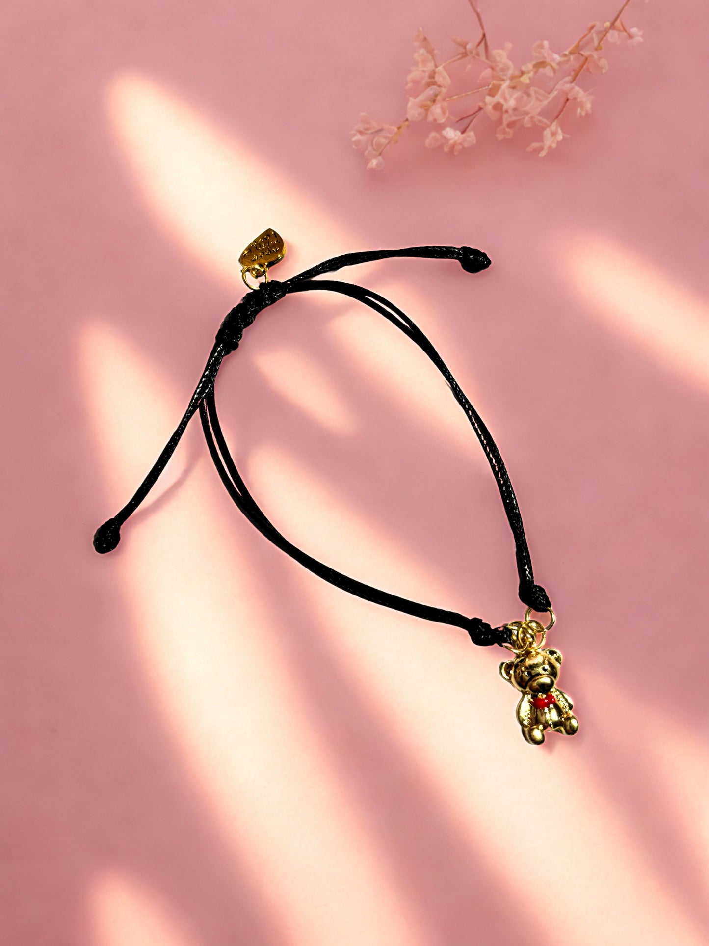 Black cord bracelet with a gold teddy bear charm on a pink background