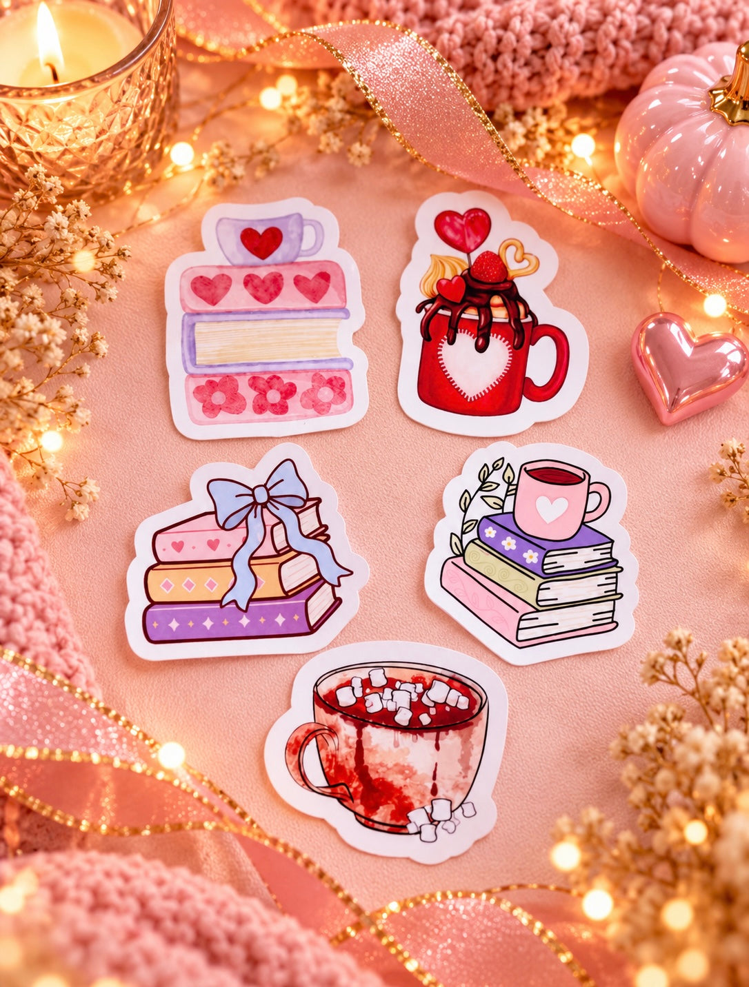 Five cute, book-and-coffee-themed die-cut stickers arranged on a pink fabric background with cozy decorative props including a candle, ribbon, fairy lights, and a small pumpkin.