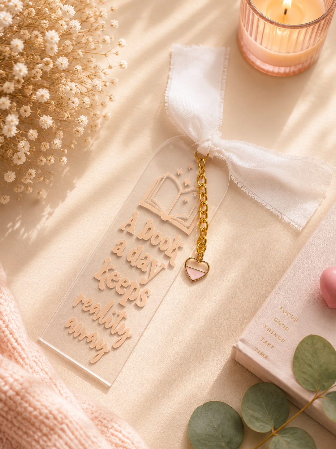 A clear acrylic book bookmark with a white ribbon bow, gold chain, and pink heart charm, featuring the text 'A book a day keeps really busy'. The bookmark is styled in a warm, cozy flat-lay setting with dried flowers, a candle, a cozy knit blanket, and a book.