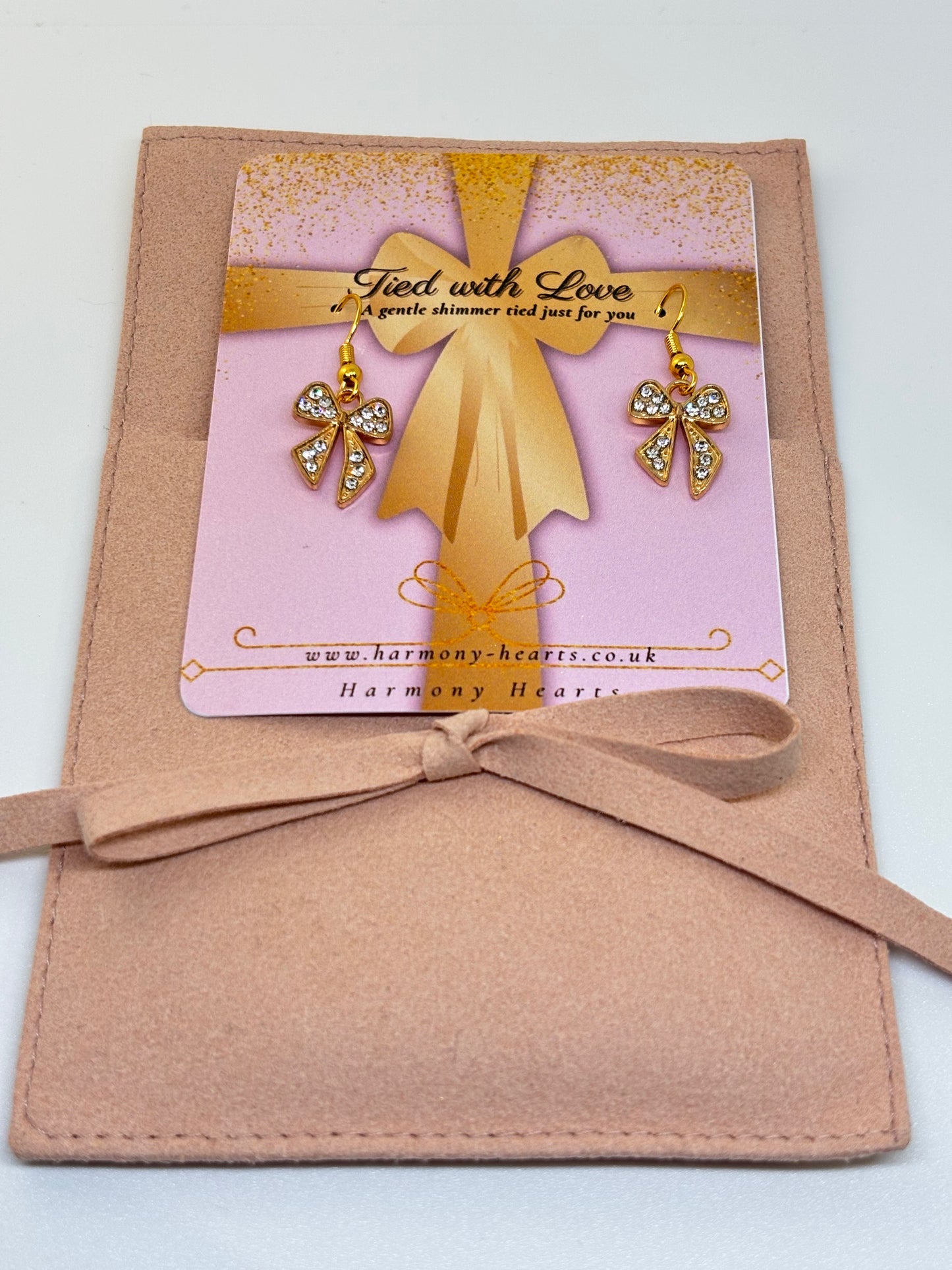Gold bow with earrings on a pink card inside a brown envelope, labeled 'Harmony Hearts'.