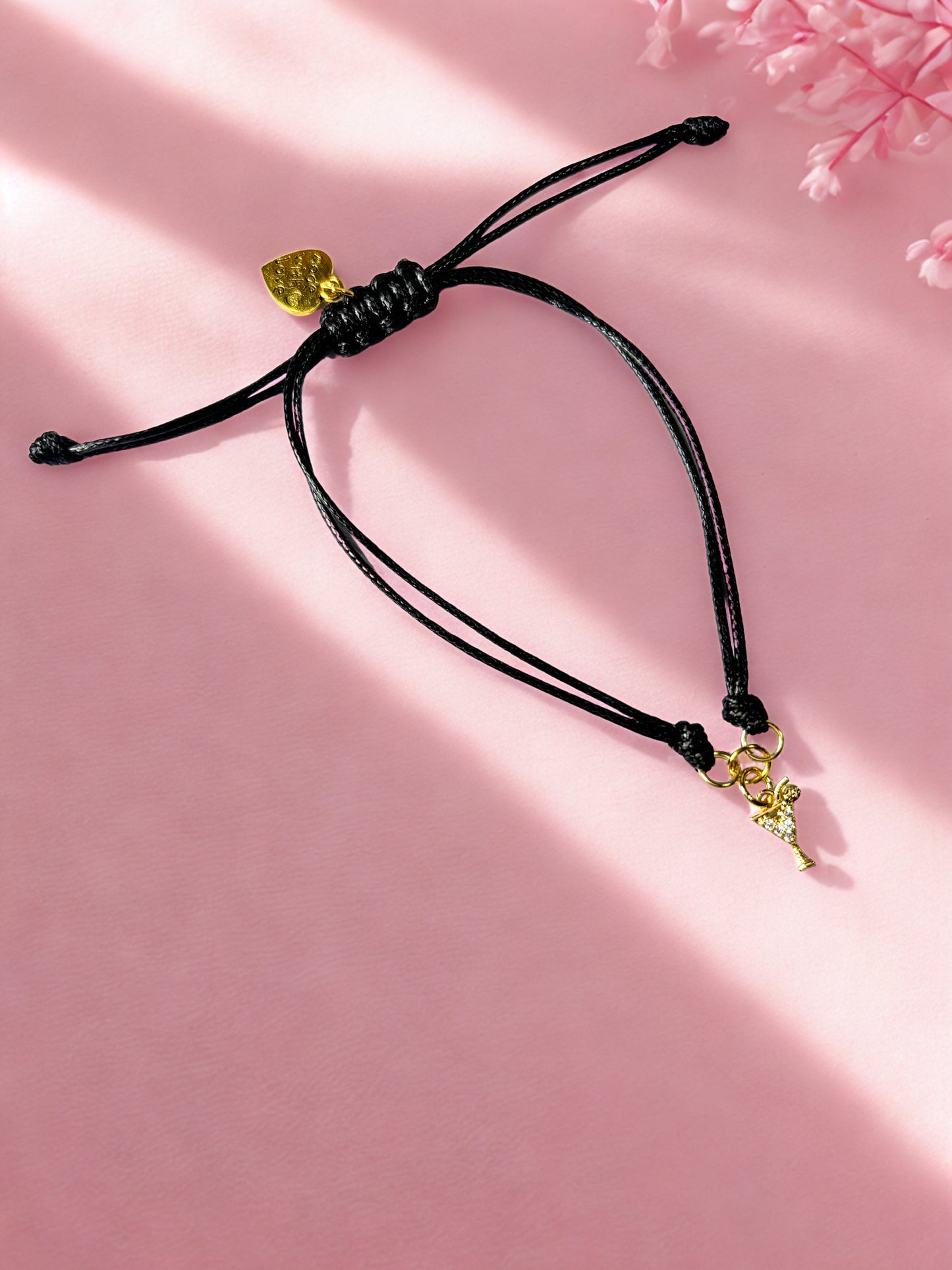 Black cord bracelet with gold cocktail charm on a pink background