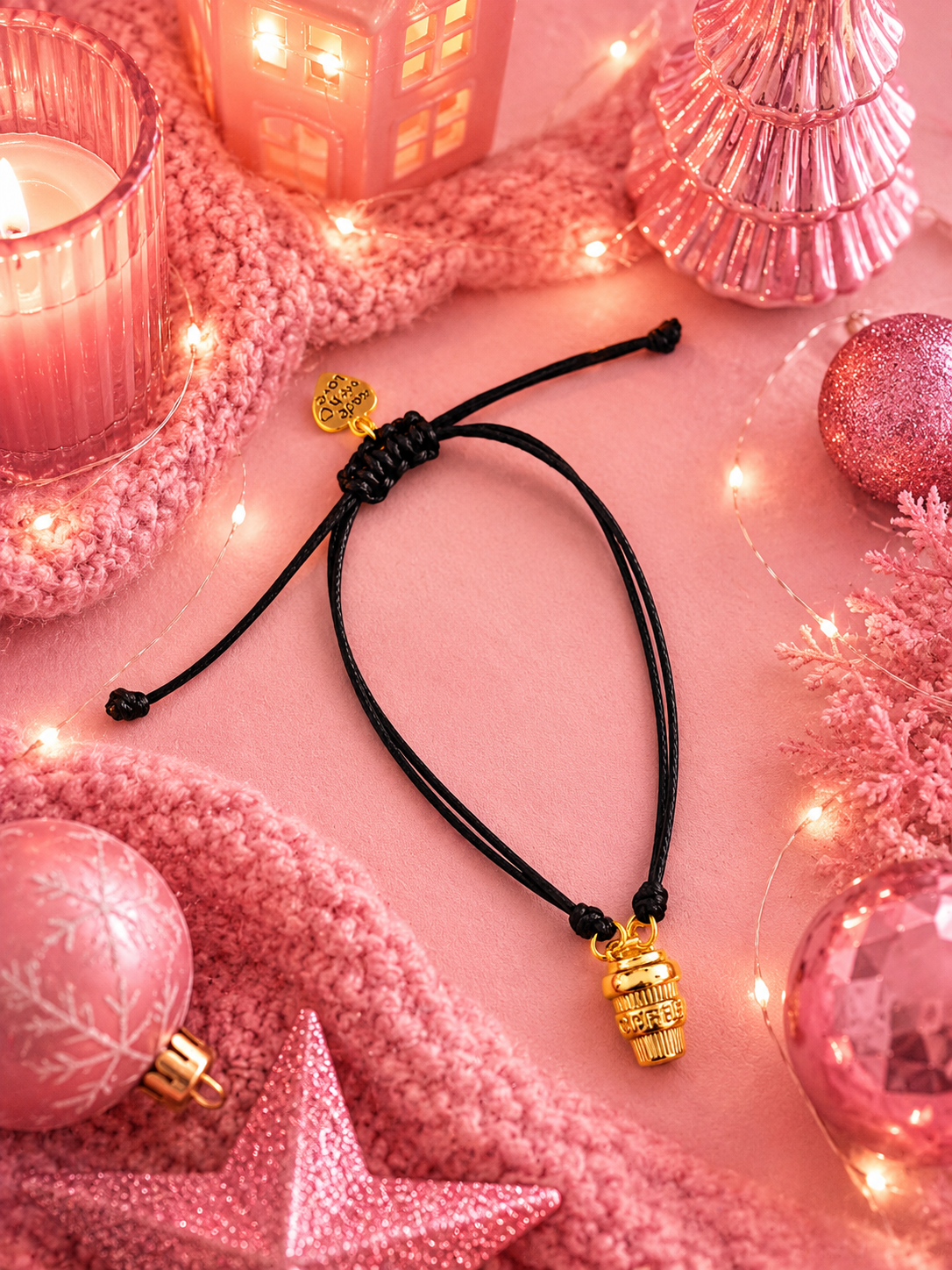 A black cord adjustable bracelet with a small gold-toned coffee charm, styled on a pink holiday-themed background with pink ornaments, a knit scarf, and warm string lights.