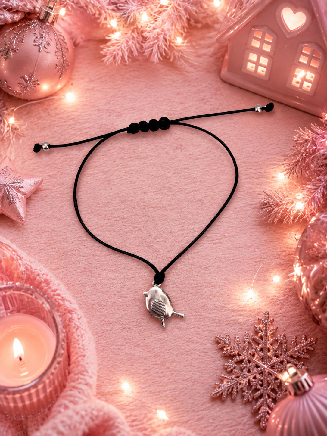 A black cord charm bracelet with a small silver robin charm, styled on a pink textured surface with warm fairy lights, pink ornaments, a candle, and a decorative house.