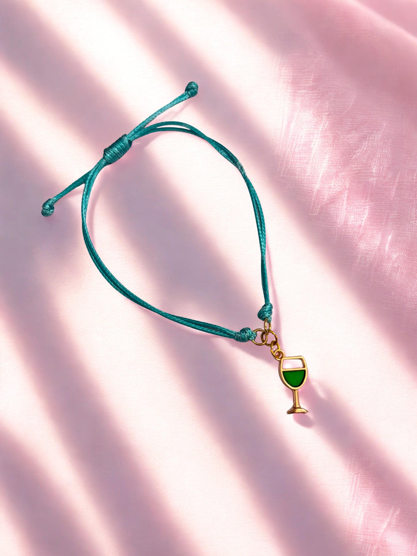 Teal cord bracelet with a green and gold wine glass charm on a pink fabric background