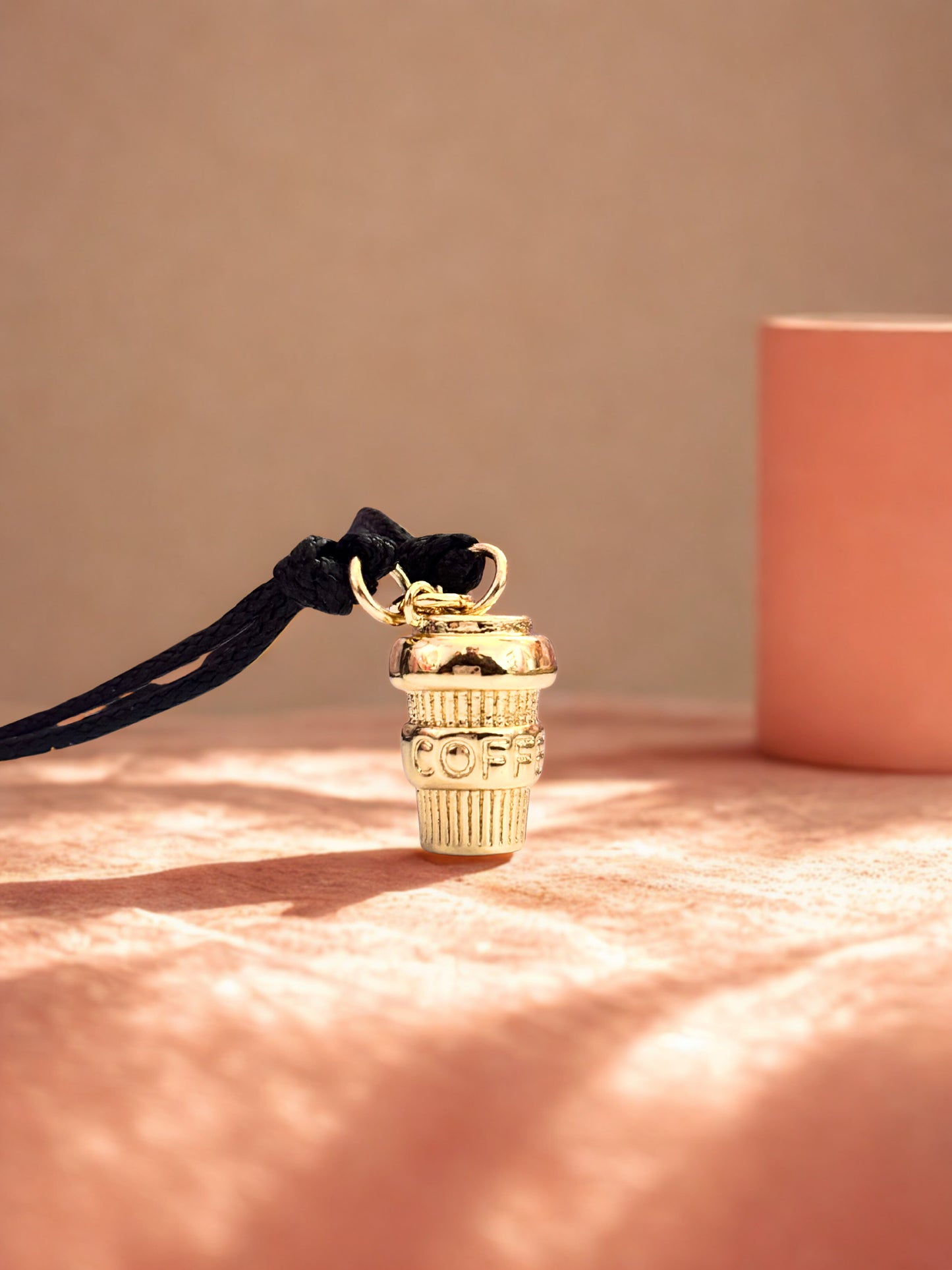 Gold coffee charm bracelet on a black cord against a pink background