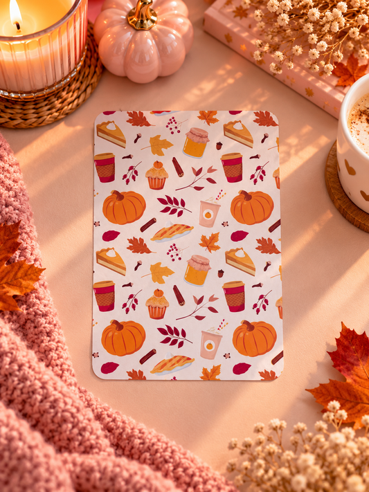 A rectangular kindle insert with an autumn-themed pattern of pumpkins, leaves, cupcakes, and coffee cups, styled on a warm beige surface with a pink knit blanket, candles, dried flowers, and a small pumpkin.