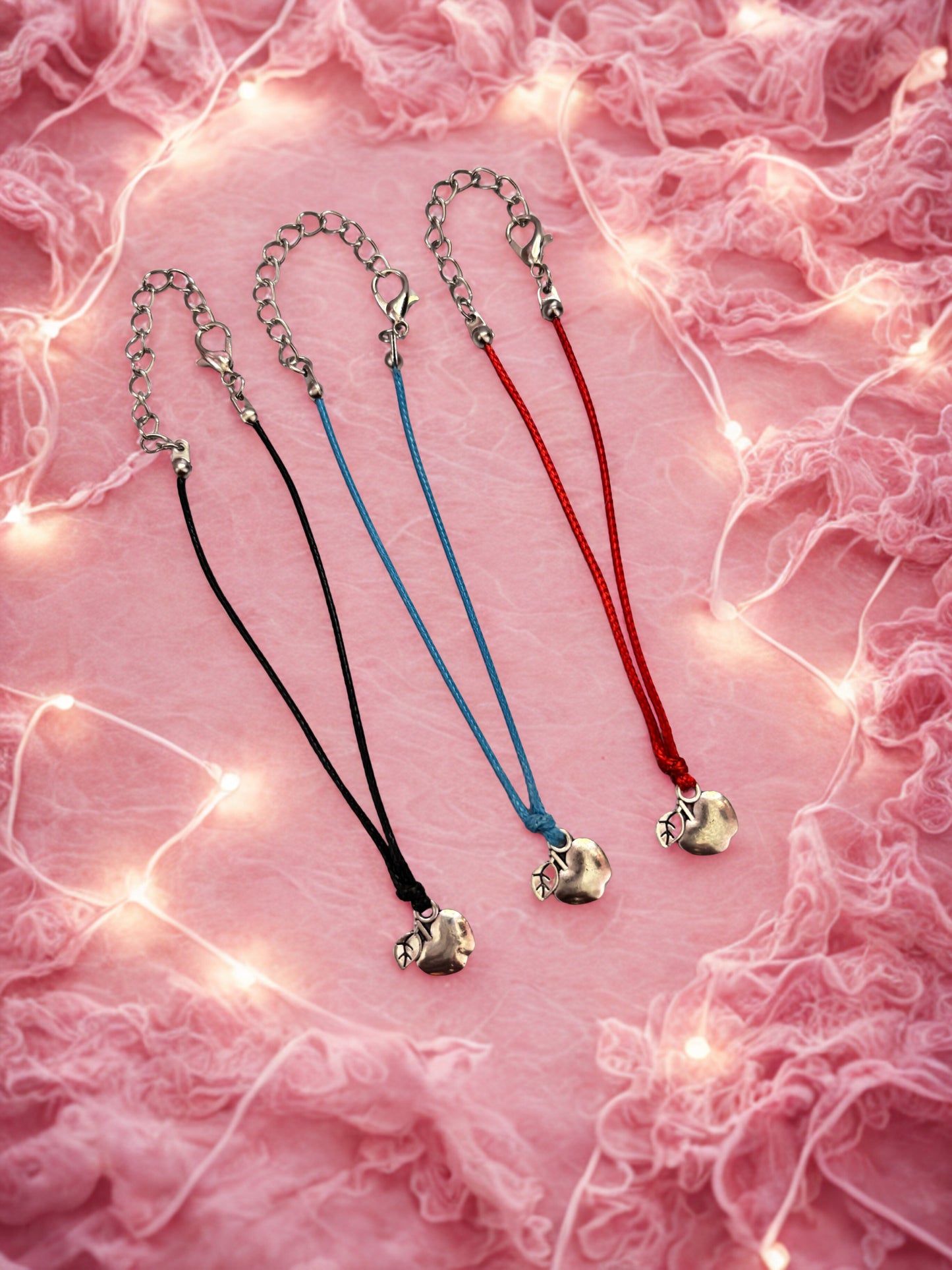 Three bracelets with apple-shaped pendants on a pink textured background with string lights.