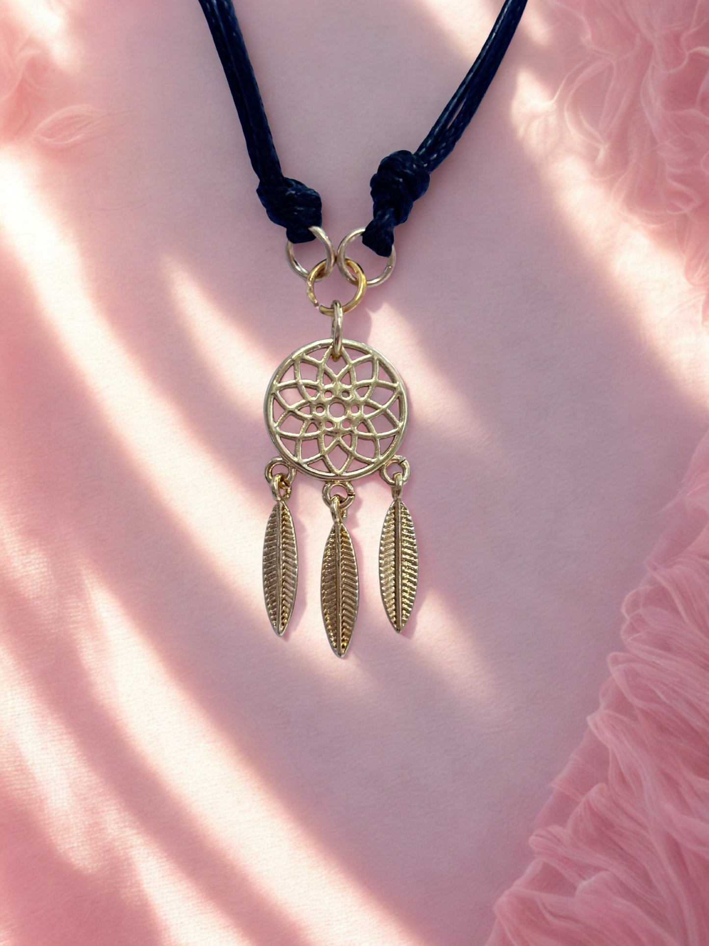 Gold dreamcatcher charm with feathers on a black cord against a pink fabric background