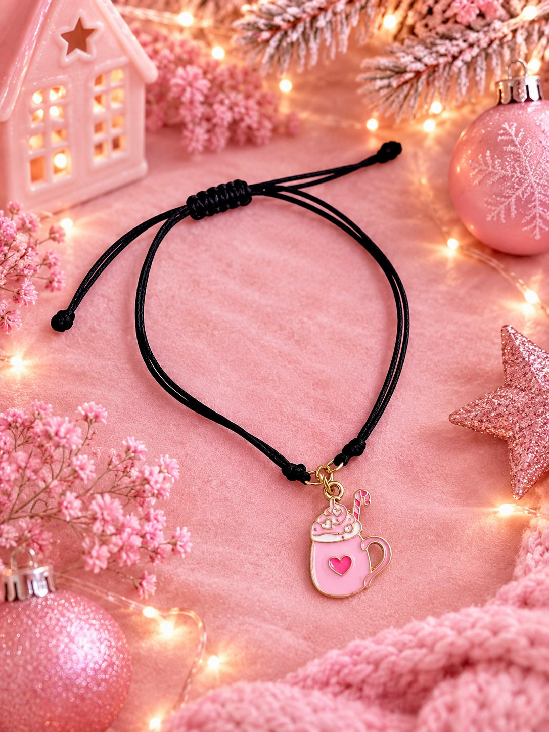 A black adjustable cord bracelet with a pink and gold hot chocolate mug charm, styled on a pink textured fabric with holiday decorations including a white house ornament, pink ornaments, and fairy lights.