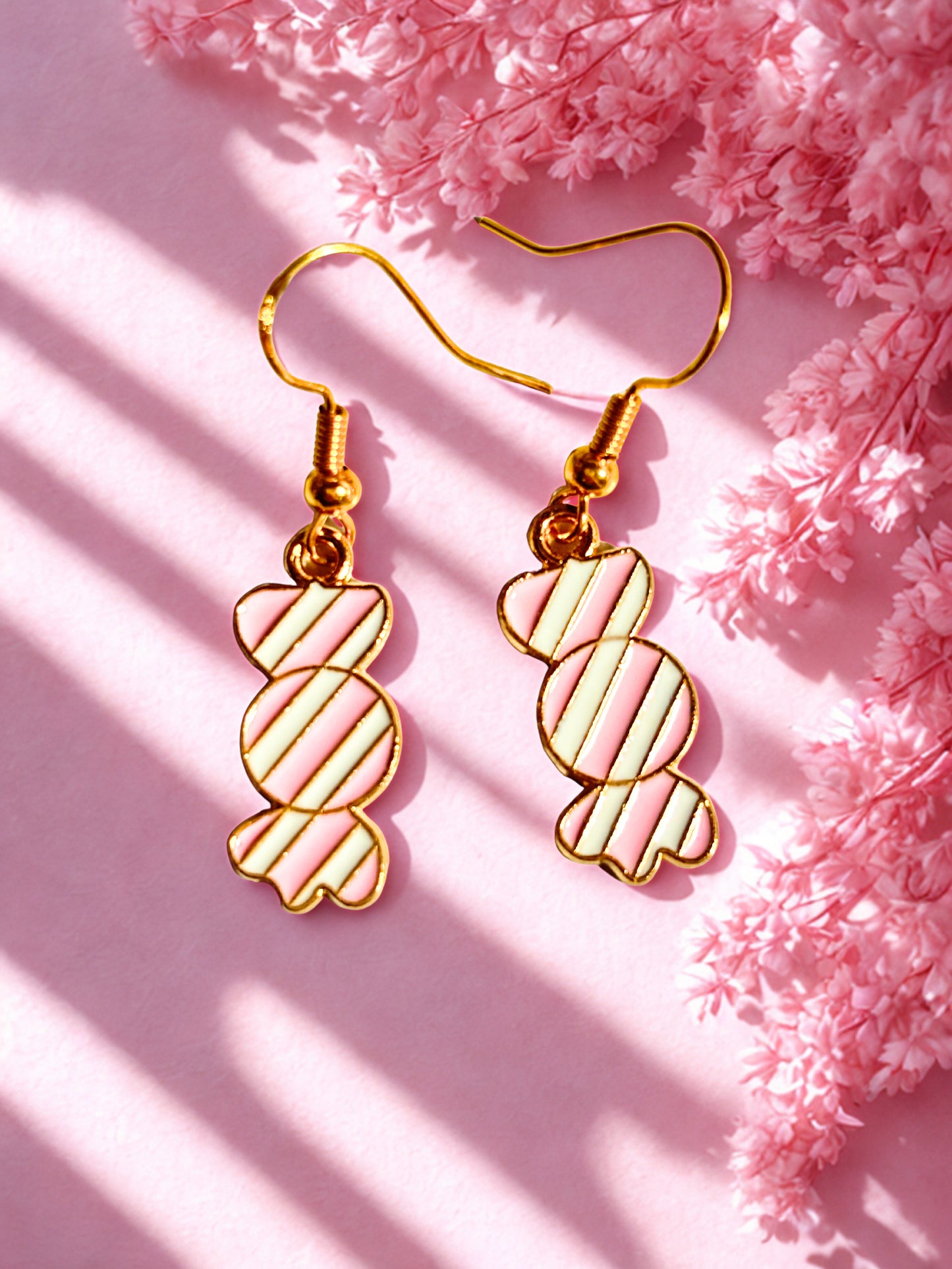 Gold pink and white candy-shaped sweet earrings on a pink background with pink flowers.