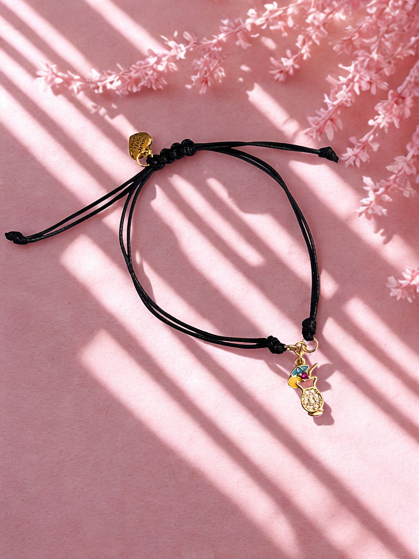 Black cord bracelet with gold cocktail charm on a pink background