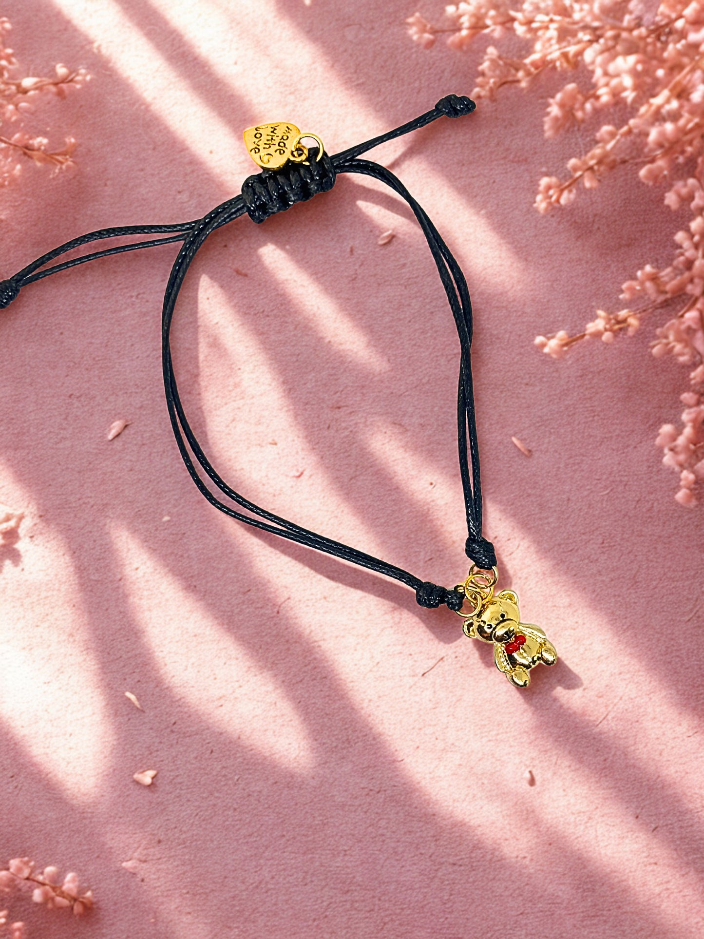 Black cord bracelet with gold Teddy Bear charm on a pink background