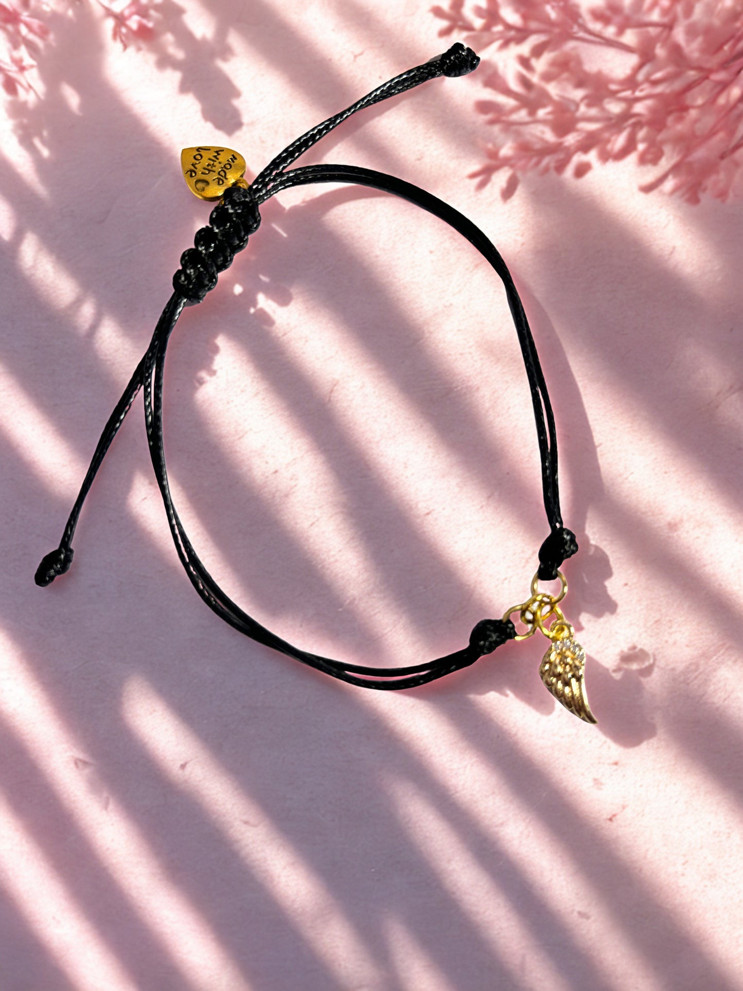 Black cord bracelet with gold angel wing charm on a pink background