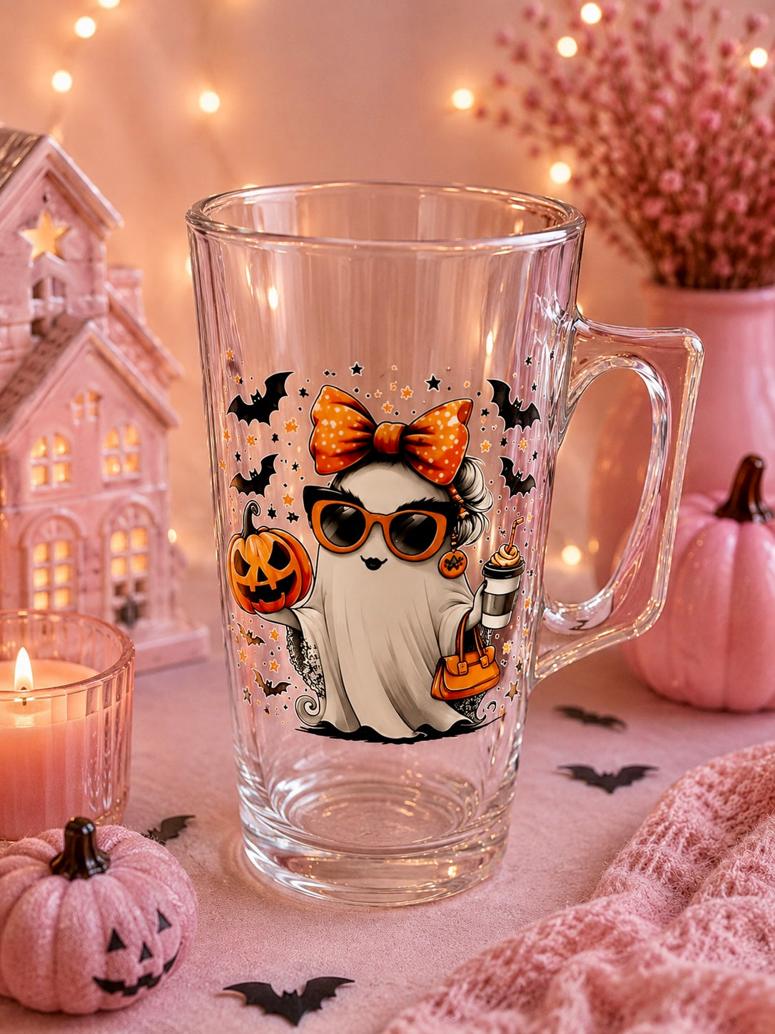 A clear glass mug with a Halloween-themed ghost illustration, set against a cozy, warm-toned background with pink pumpkins, a lit candle, and fairy lights.