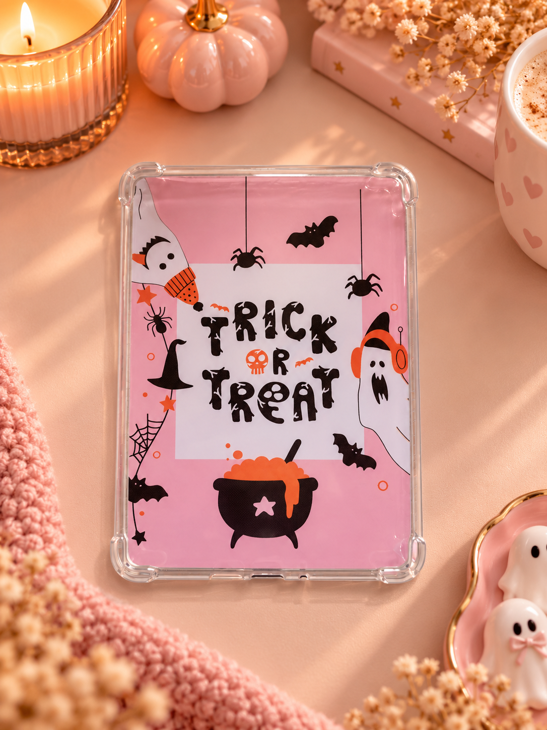 A clear plastic kindle case with a pink Halloween-themed design insert featuring the words 'TRICK OR TREAT' in black, along with ghosts, bats, a witch hat, and a cauldron, styled on a pink surface with candles and pumpkins.