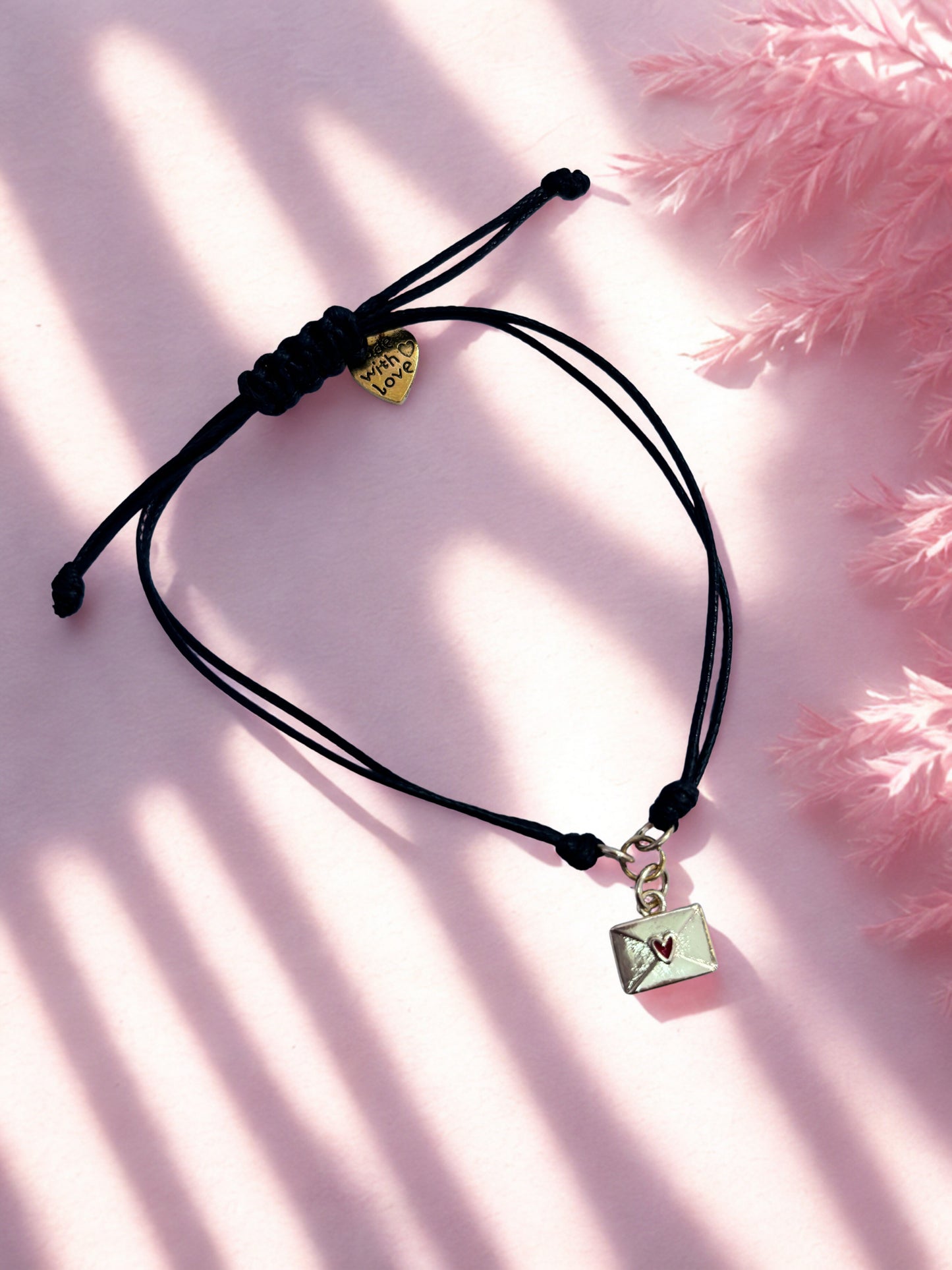 Black cord bracelet with a gold envelope charm on a pink background
