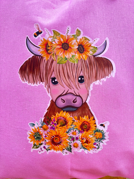 A pink fabric tote bag, featuring a printed illustration of a cute Highland cow with a sunflower crown and floral collar.
