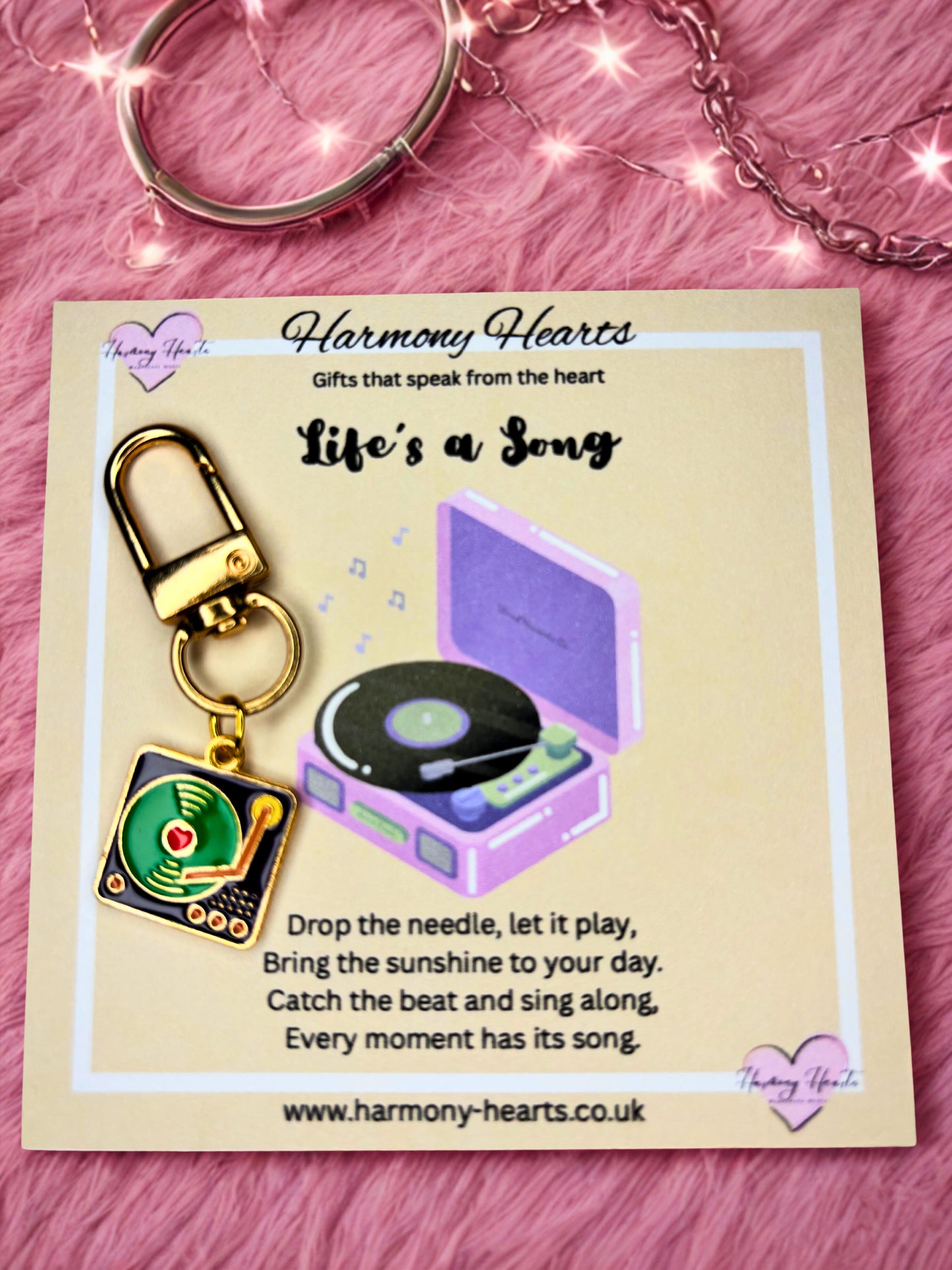 Keychain and record player charm on a 'Harmony Hearts' card with text on a pink textured background.