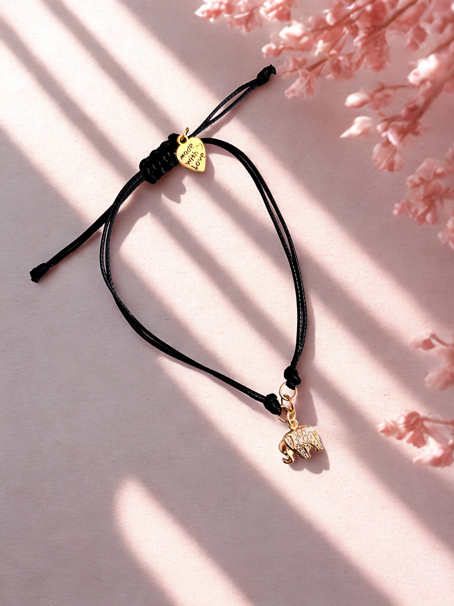 Black cord bracelet with gold elephant charm on a pink background