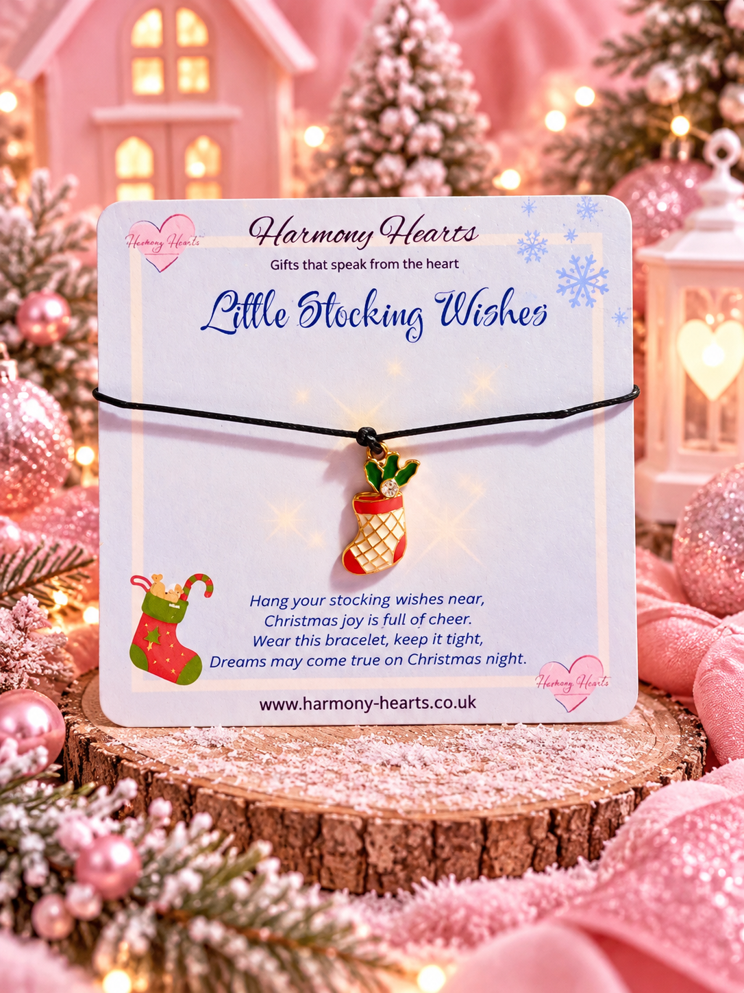 A small Christmas stocking bracelet by Harmony Hearts on a white display card, styled on a wooden slice with pink holiday decorations and warm lights in the background.