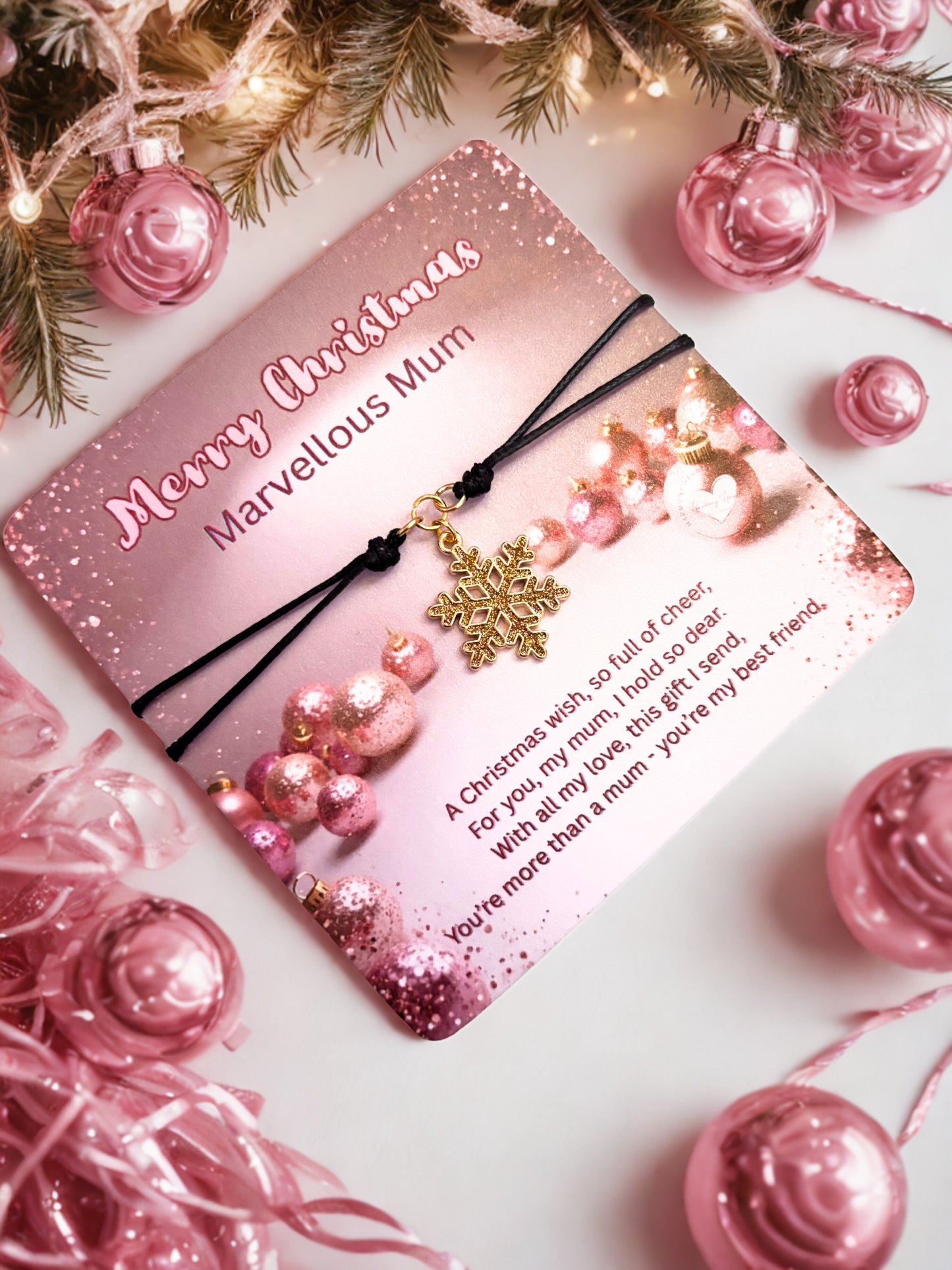 Pink Christmas card with gold snowflake charm bracelet surrounded by pink ornaments and tree branches.