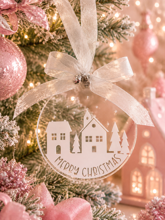 A round clear acrylic Christmas ornament with a white silhouette of houses and trees and the words 'Merry Christmas' written across the bottom, hanging from a white ribbon on a decorated Christmas tree.