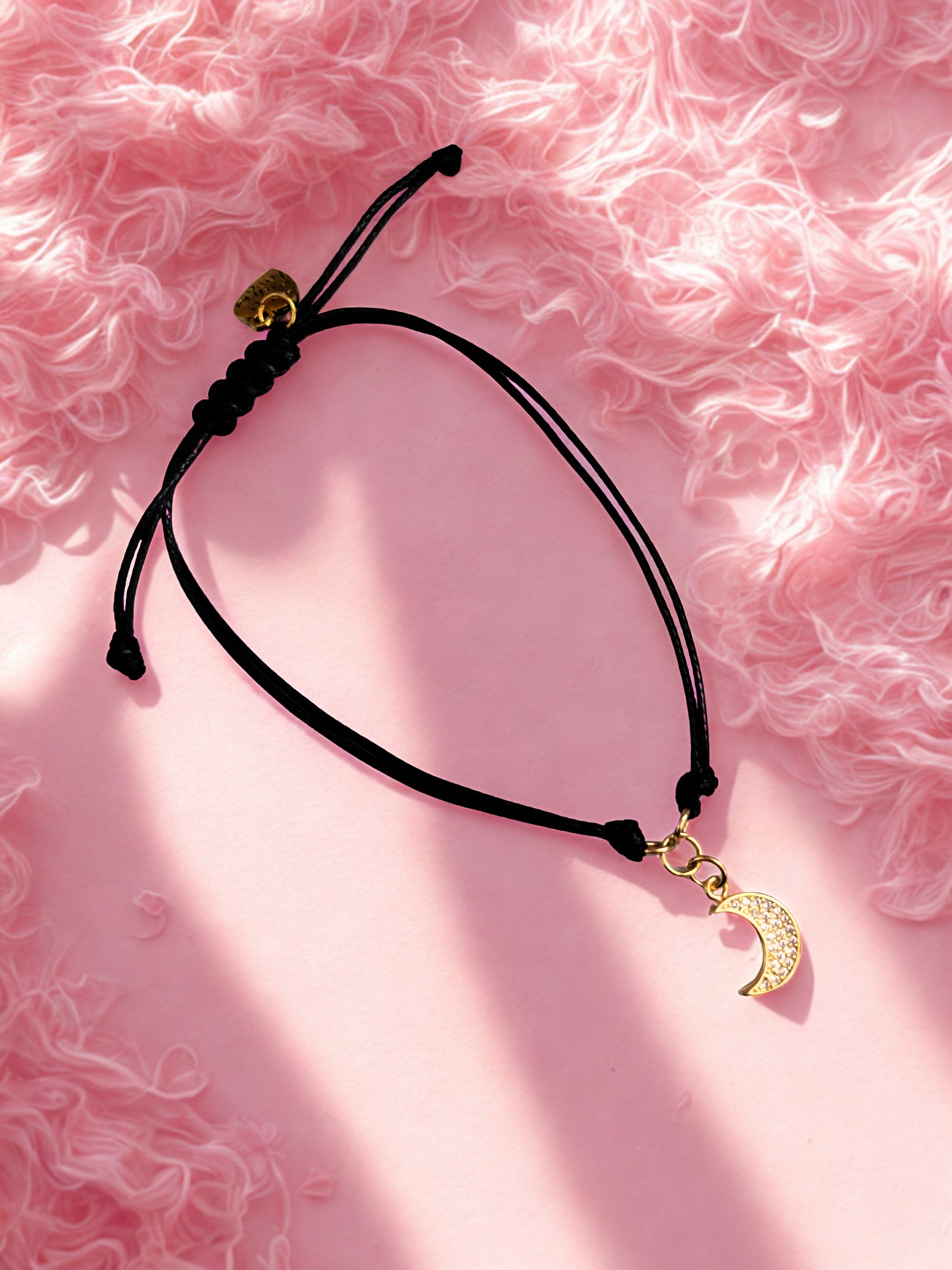 Black cord bracelet with gold crescent moon charm on a pink textured background