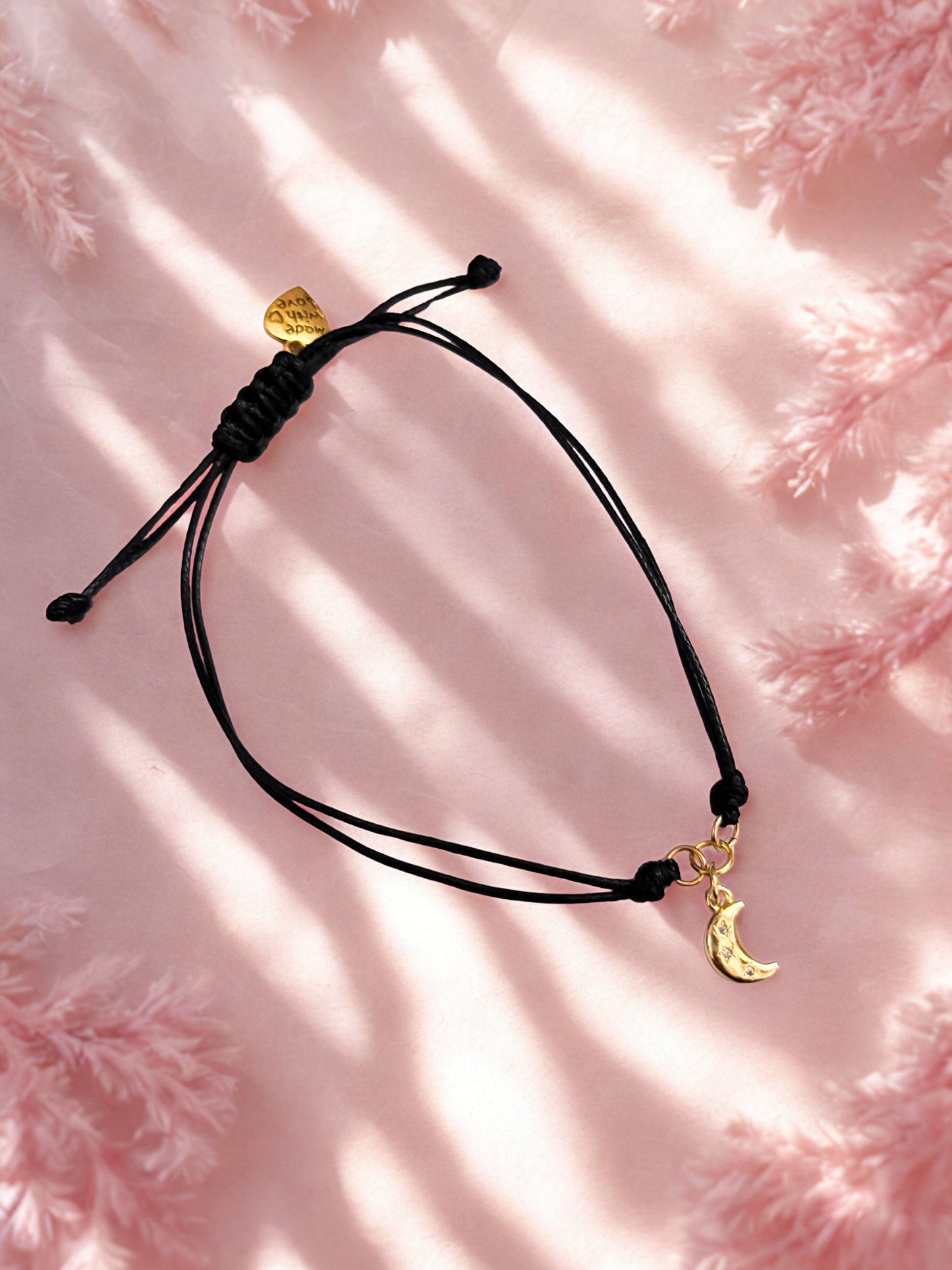 Black cord bracelet with gold moon charm on a pink textured background