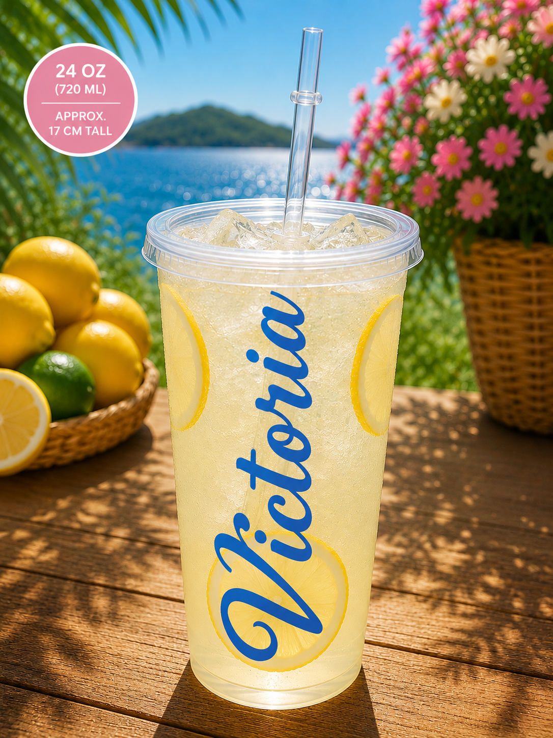 A clear 24 oz plastic tumbler cup with a clear straw, filled with a pale yellow iced drink and printed with the name 'Victoria' in blue script over yellow lemon graphics. The cup sits on a wooden table outdoors with lemons, flowers, and a lake in the background.