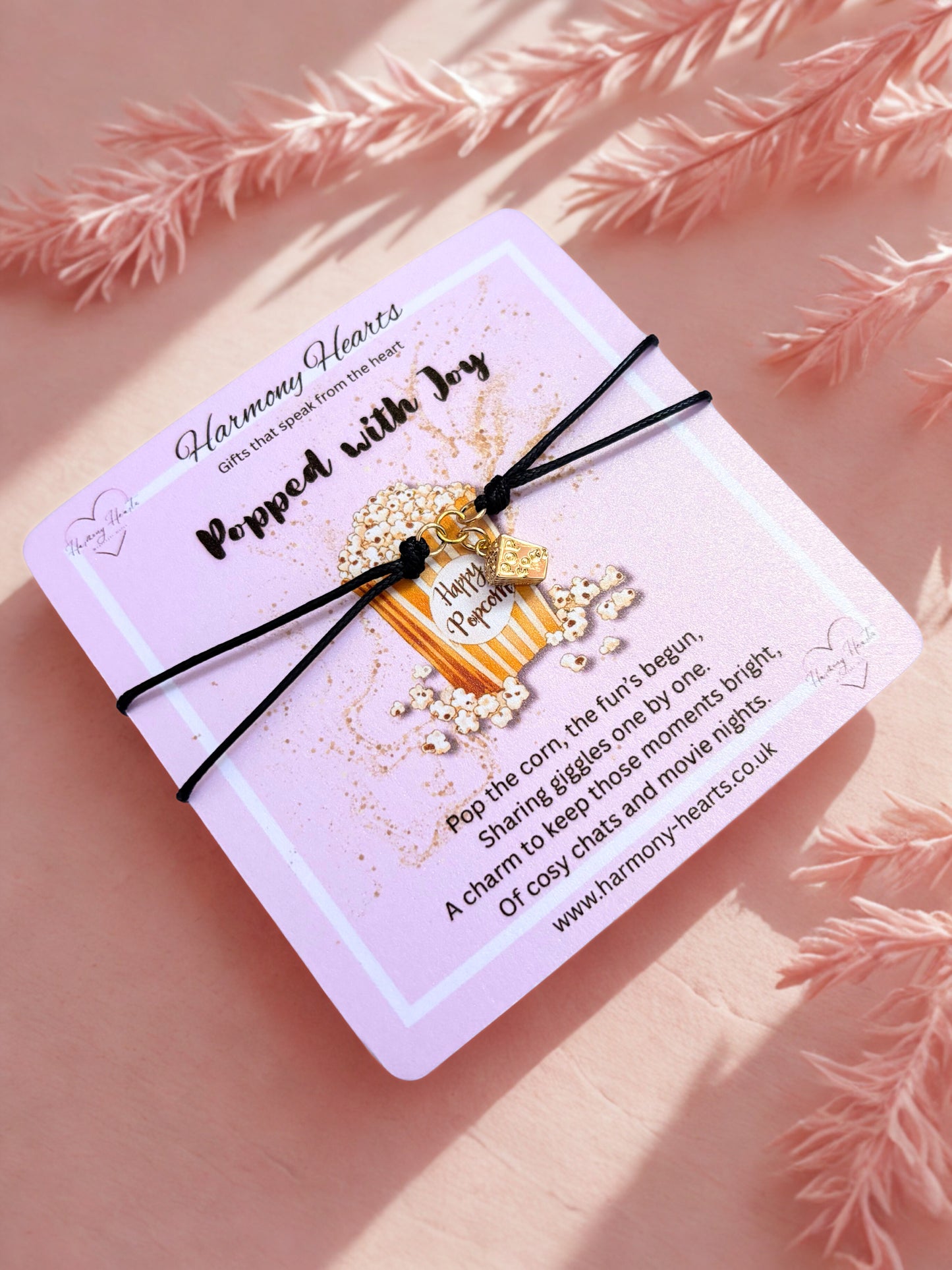 Decorative popcorn charm bracelet on a card with text, placed on a pink surface with decorative elements.