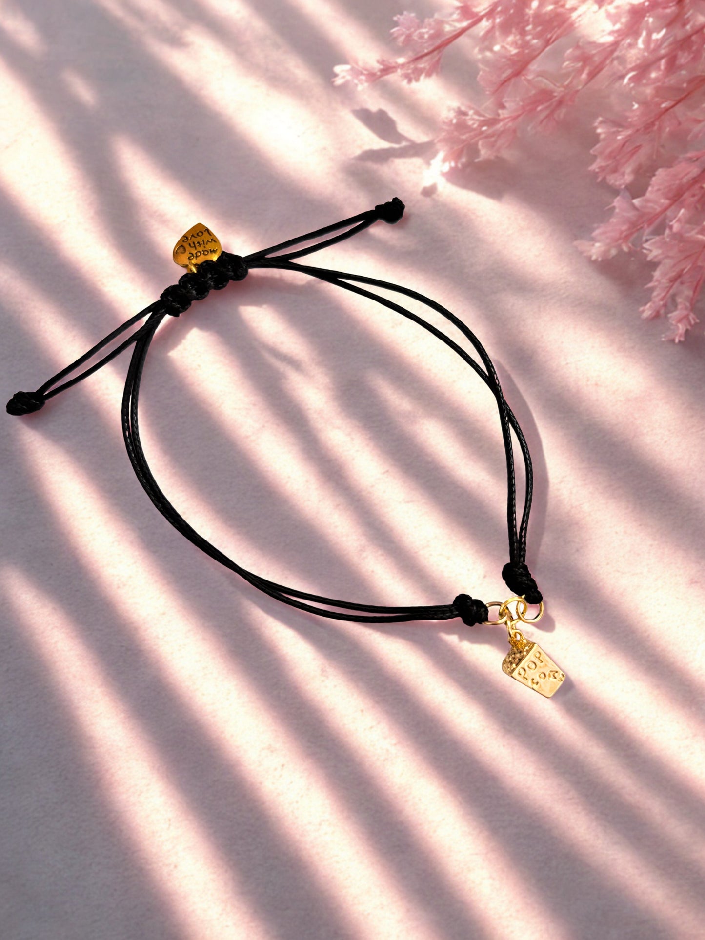 Black cord bracelet with a gold popcorn charm on a soft pink background