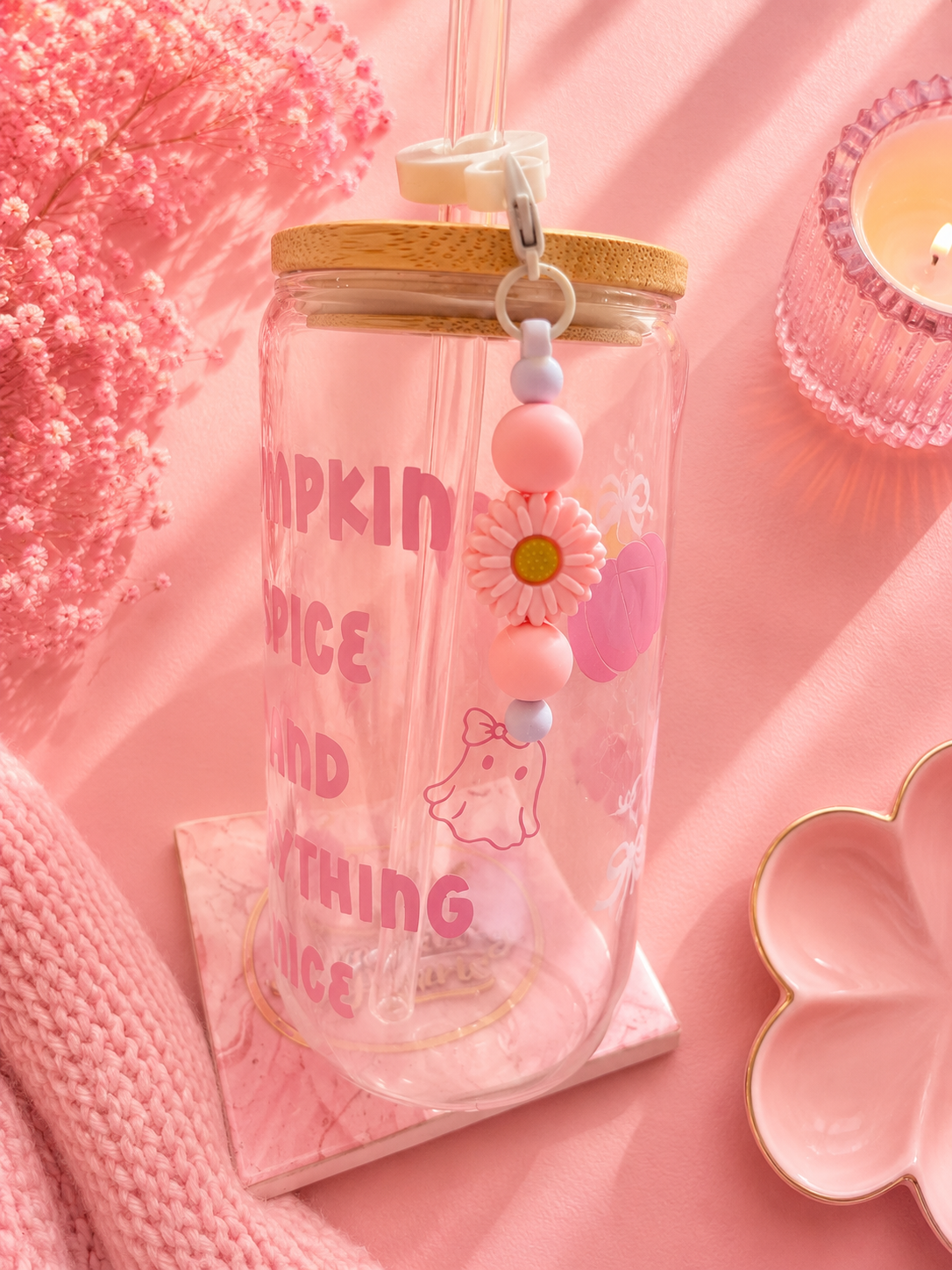 A clear glass mason jar tumbler with a bamboo lid, decorated with pink text reading 'PUMPKIN SPICE AND EVERYTHING NICE', a pink ghost illustration, and a dangling pink flower charm. It is styled on a pink fabric background with dried flowers, a candle, and a pink knit blanket.