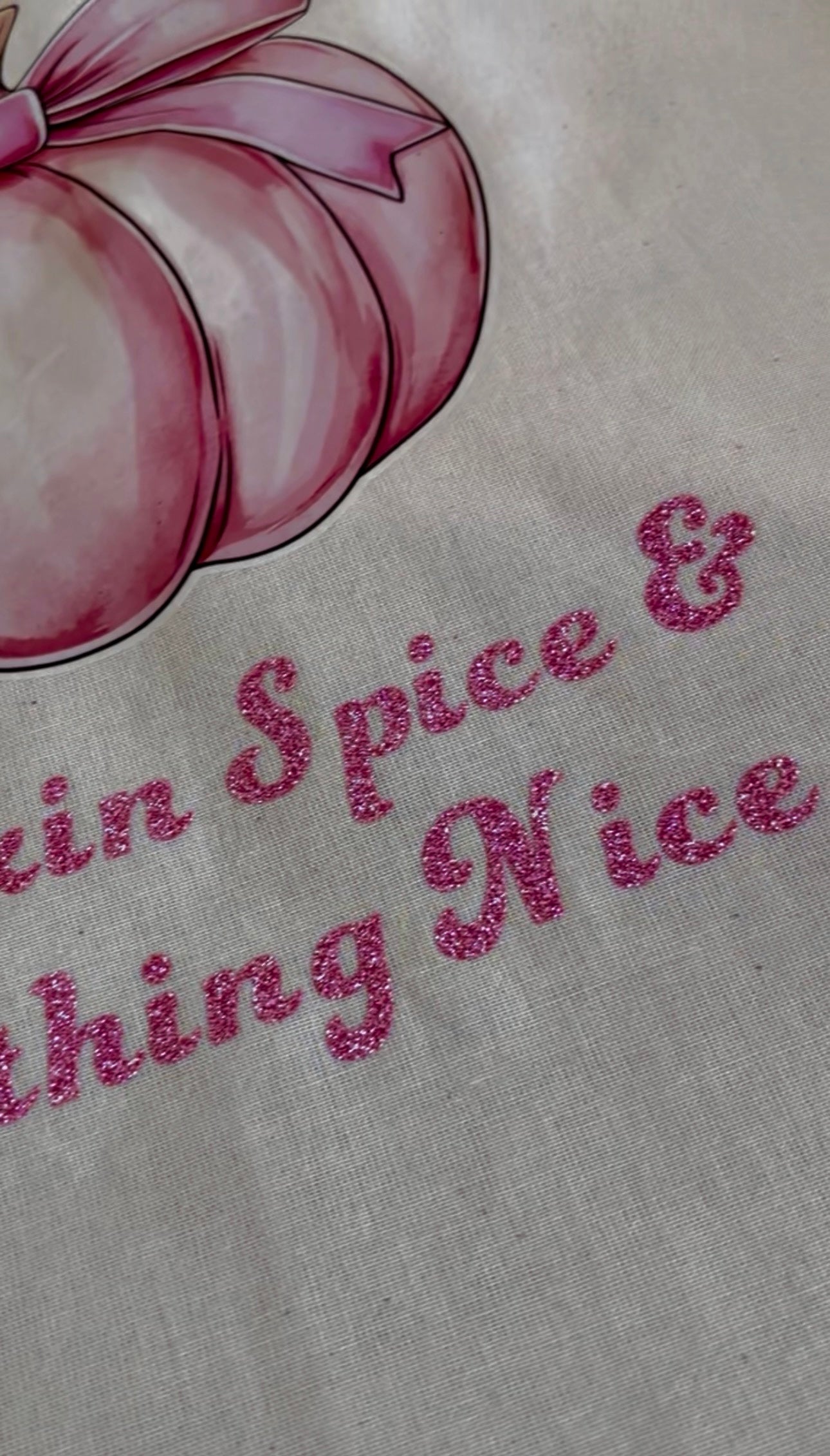 Close-up of a light beige fabric surface with a pink illustrated pumpkin and pink glitter-style text reading 'Skin Spice & Thing Nice'.