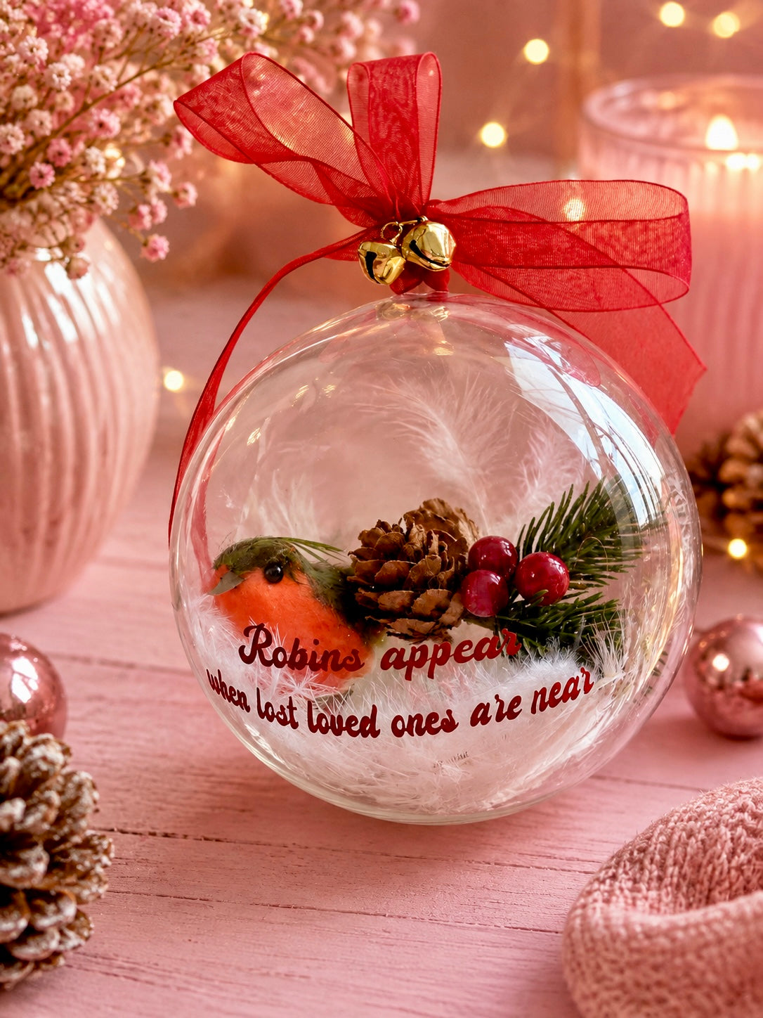 A clear round Christmas ornament with a red ribbon bow and gold jingle bells, decorated with a robin, pinecone, evergreen branch, and red berries. The ornament has the phrase 'Robins appear when lost loved ones are near' printed on it.
