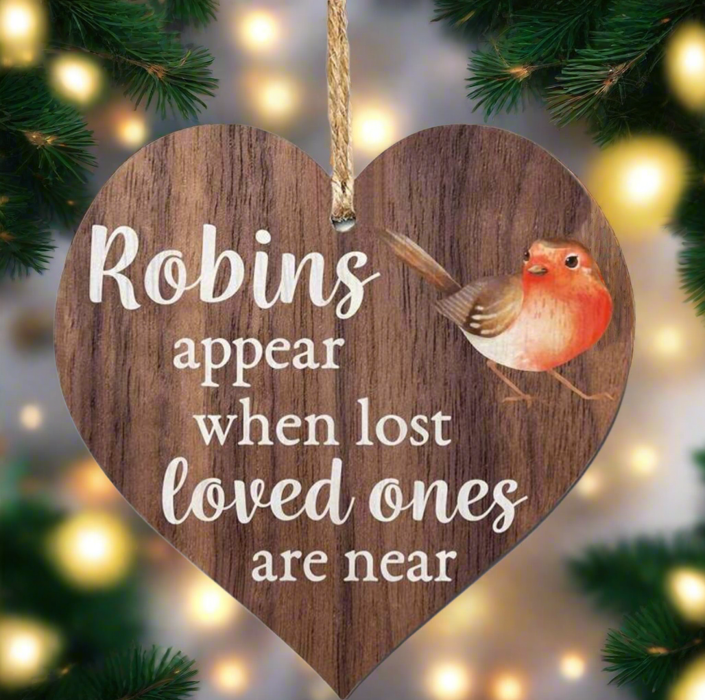 Wooden heart-shaped ornament with a robin illustration and text, hanging against a festive background with lights and greenery.