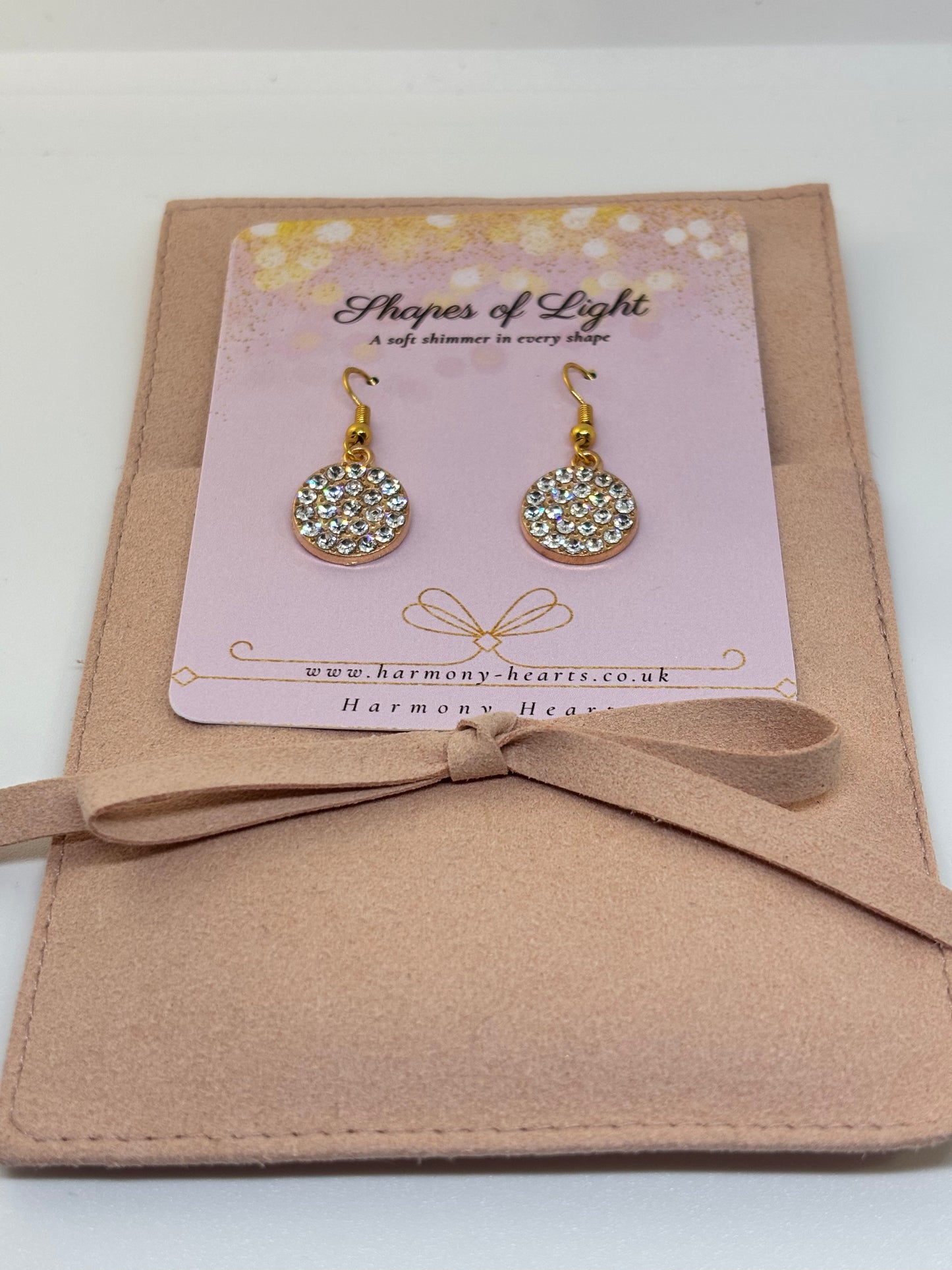 Gold earrings with crystal embellishments on a 'Shapes of Light' branded card, set against a beige background.