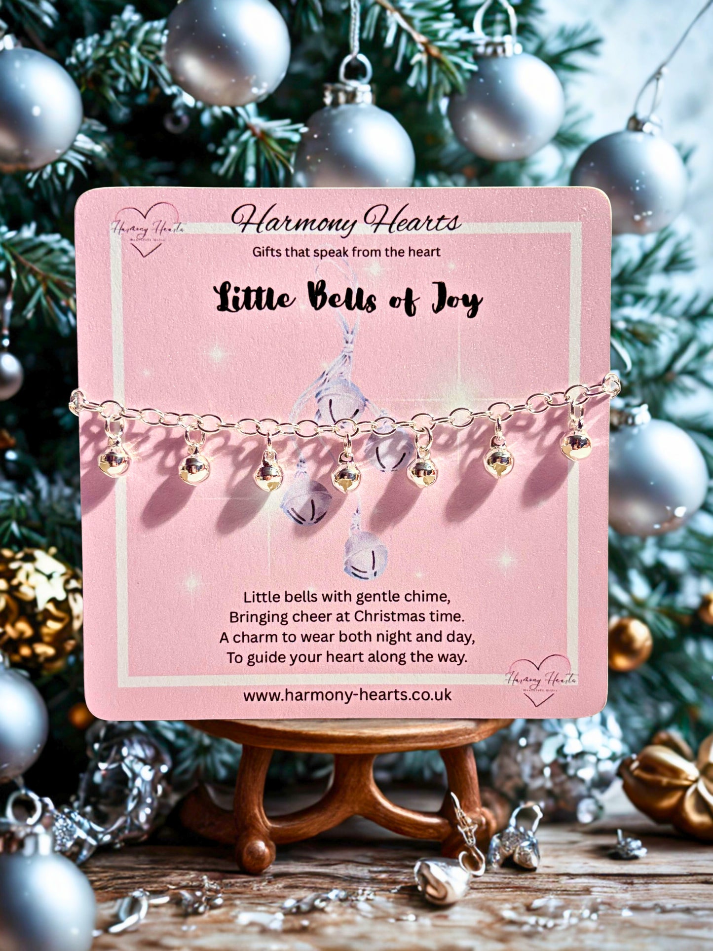 Pink card with 'Little Bells of Joy' jewelry against a Christmas tree background