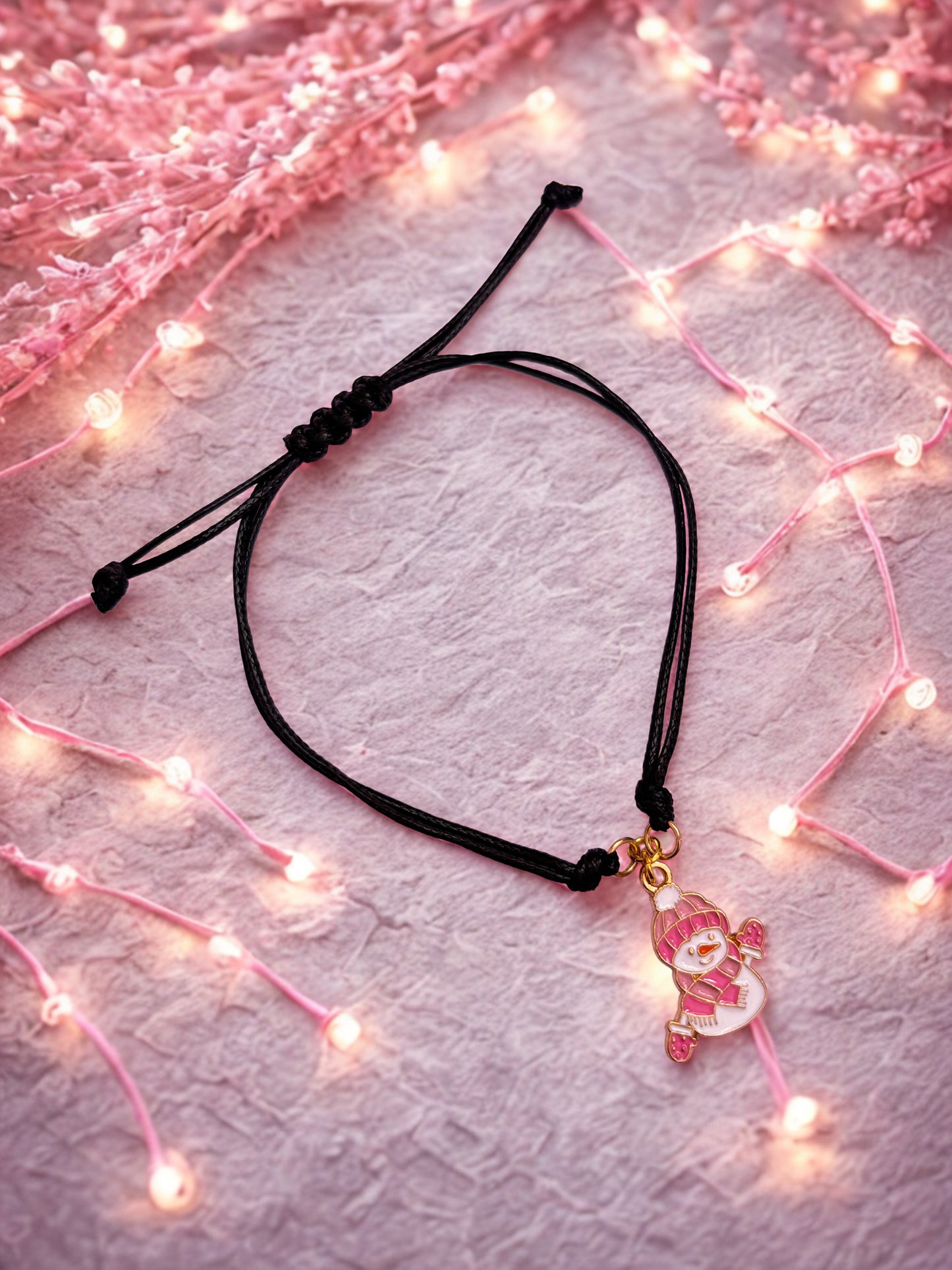 Black cord bracelet with a pink snowman charm on a light pink background with string lights.