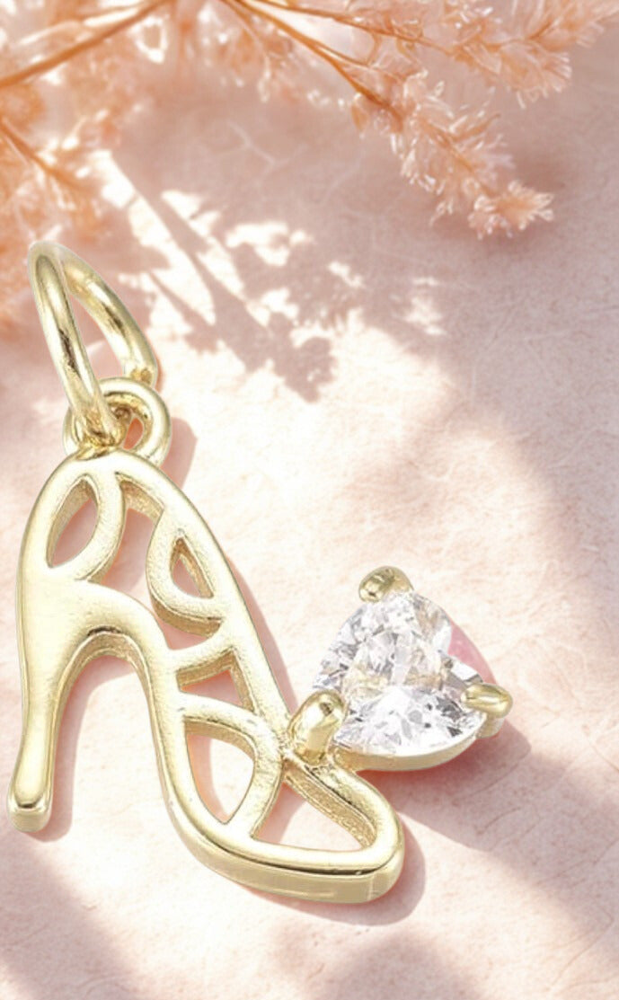 Gold high-heeled shoe charm with a clear gemstone on a soft pink background
