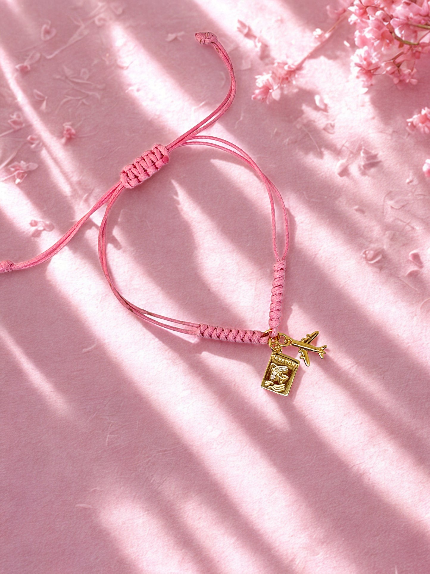 Pink braided bracelet with a gold airplane charm and passport charm on a pink background