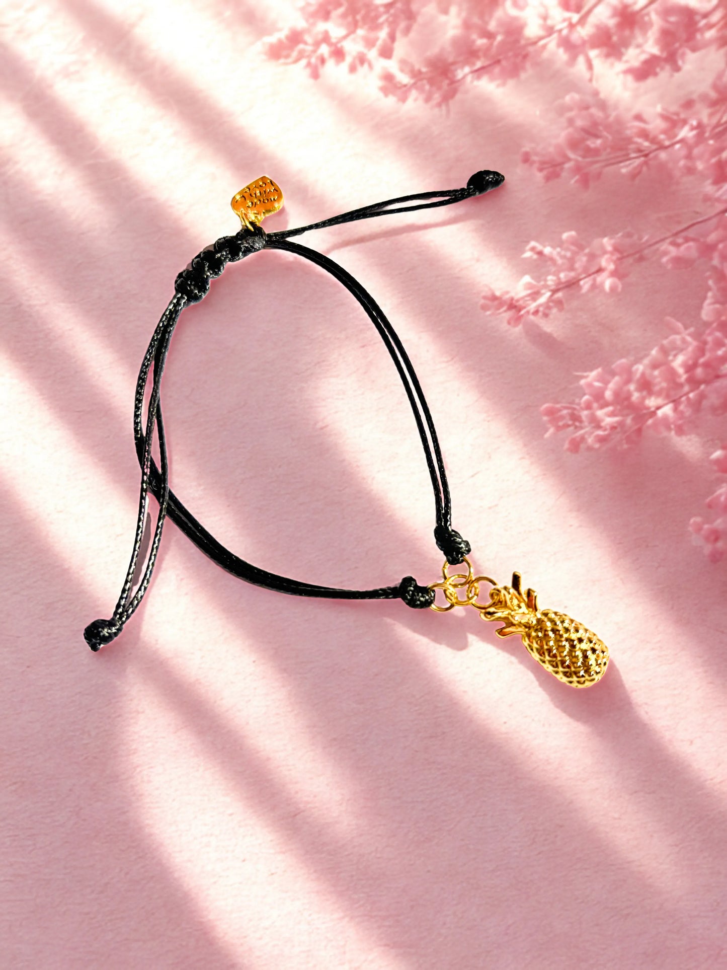 Black cord bracelet with gold pineapple charm on a pink background