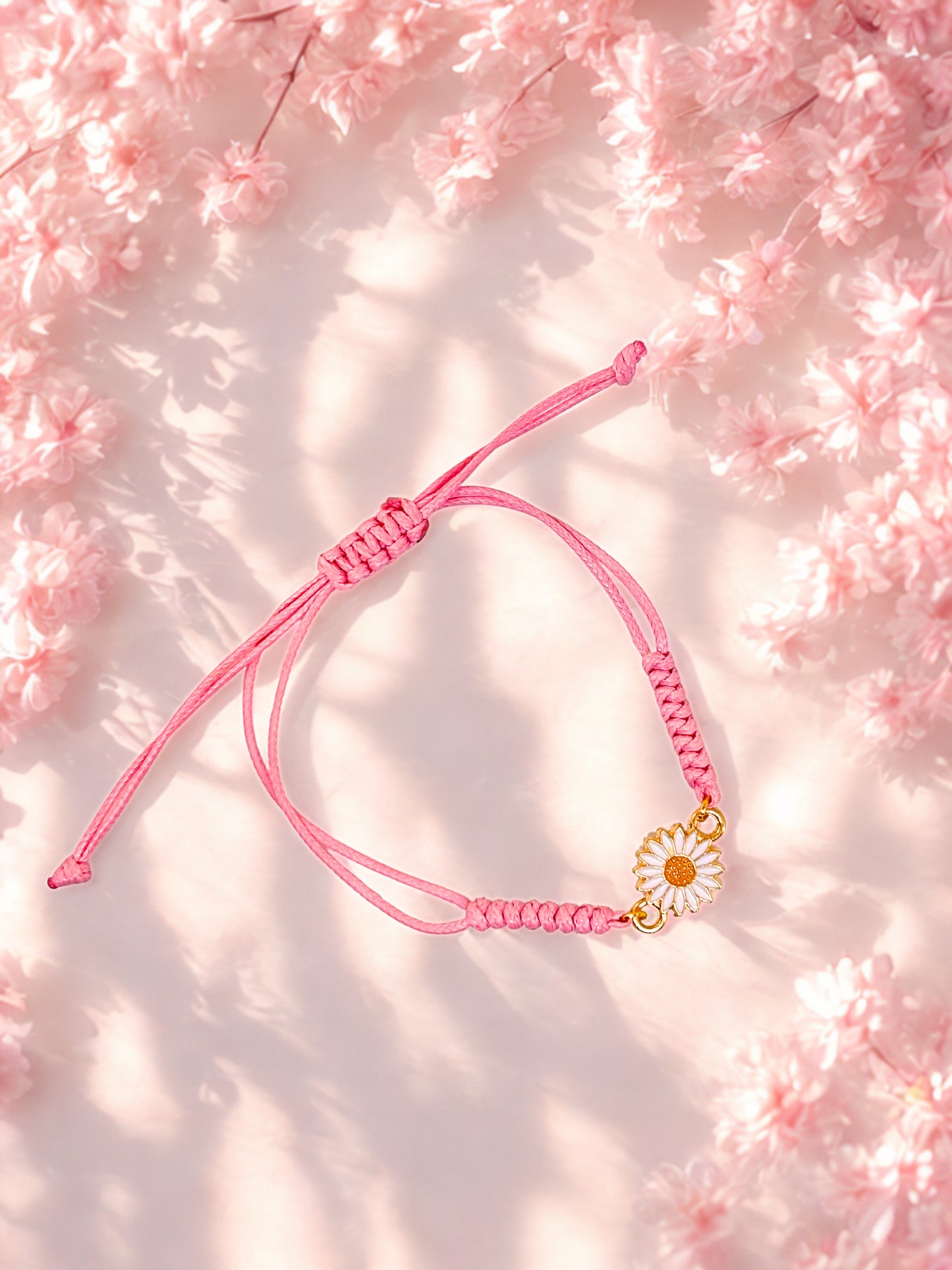 Pink braided bracelet with a gold and white daisy charm on a soft pink floral background