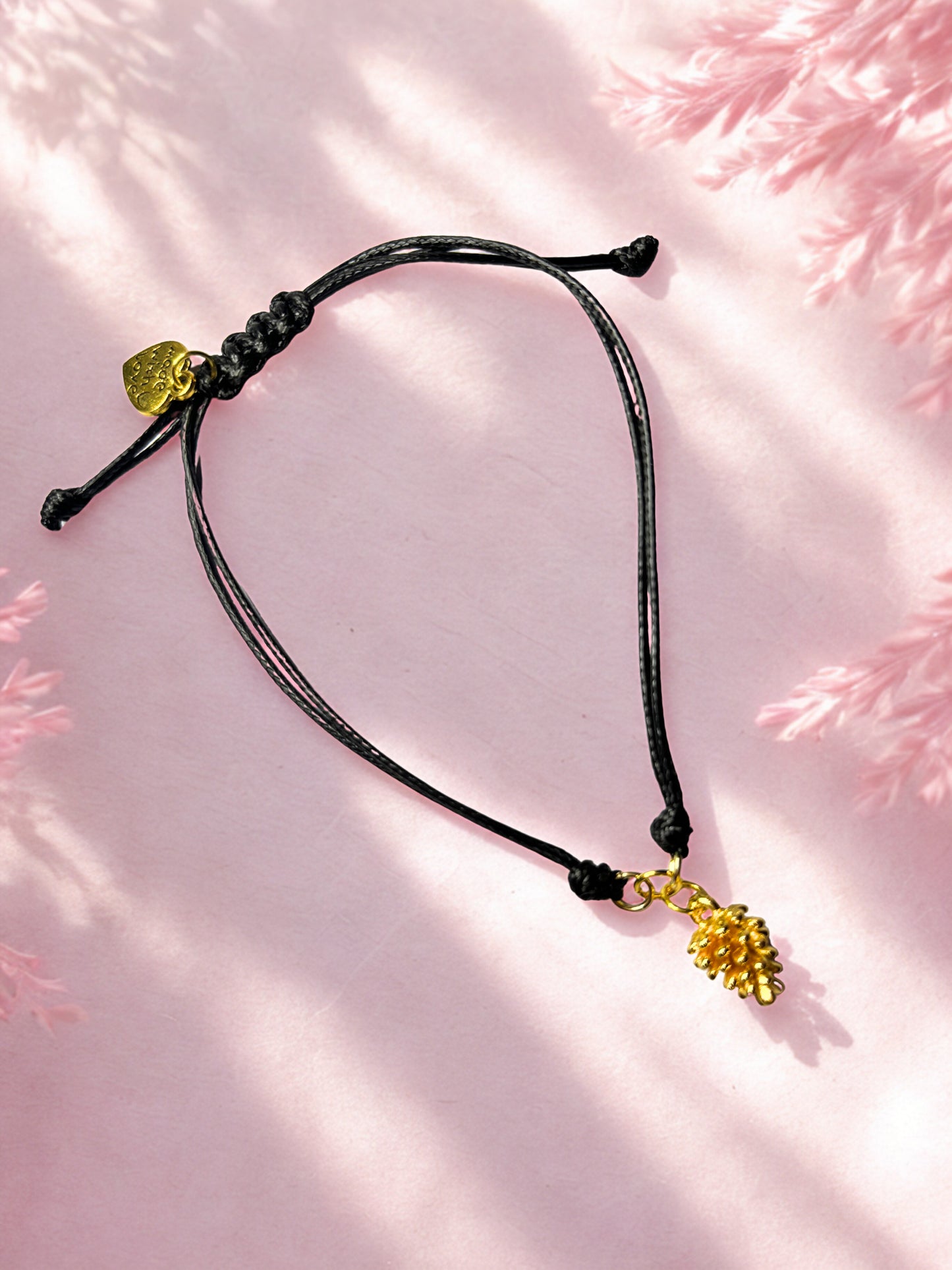 Black cord bracelet with gold pine cone charm on a pink textured background