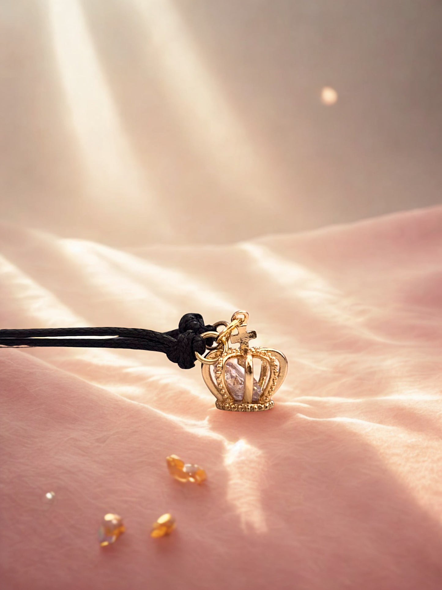 Gold crown charm on a black cord against a soft pink background
