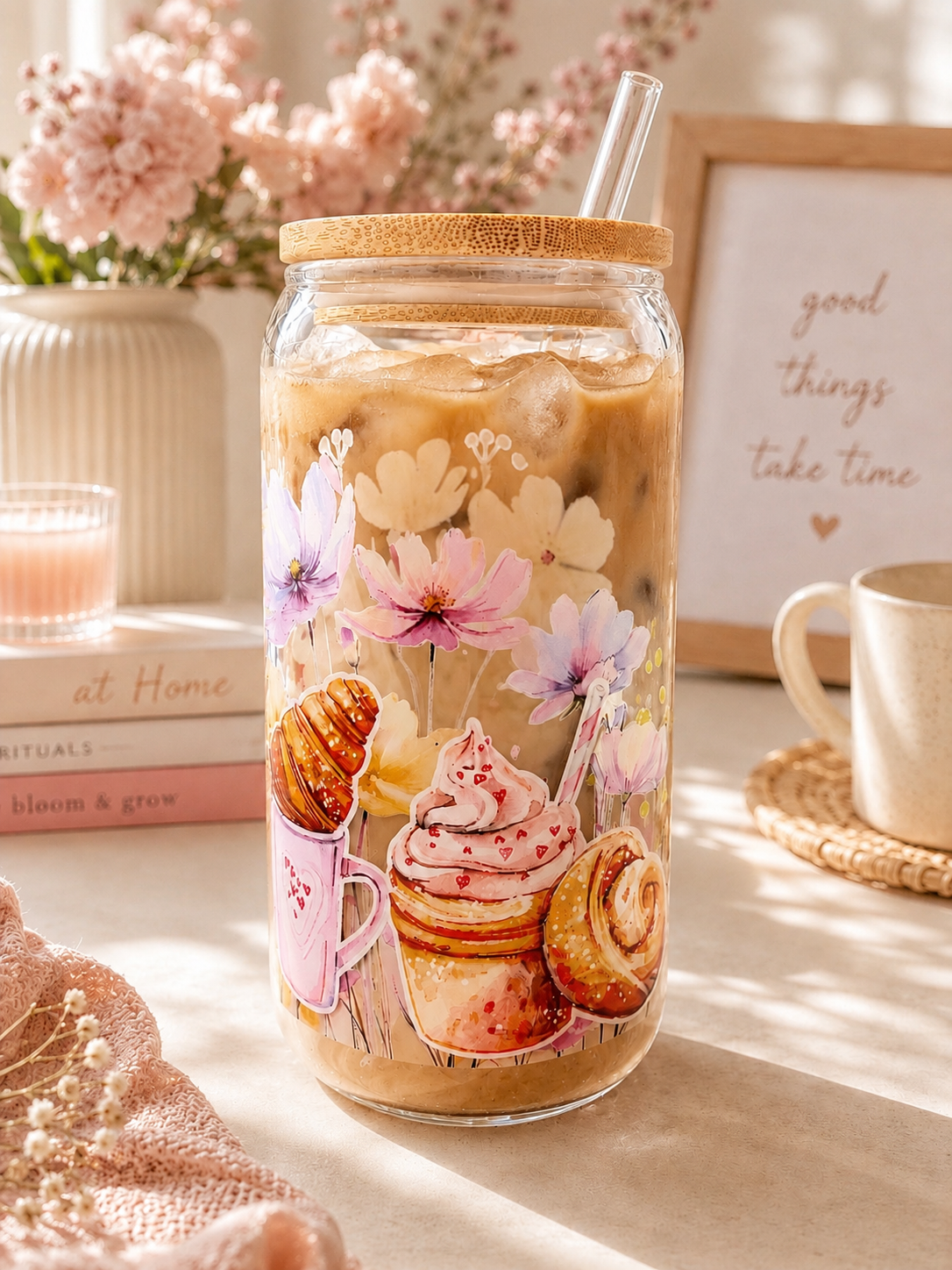 A clear glass cup with a bamboo lid and clear straw, filled with iced coffee and decorated with pastel illustrations of flowers and pastries. The jar is styled on a tabletop with a mug, books, and a framed sign in a warm, sunlit setting.