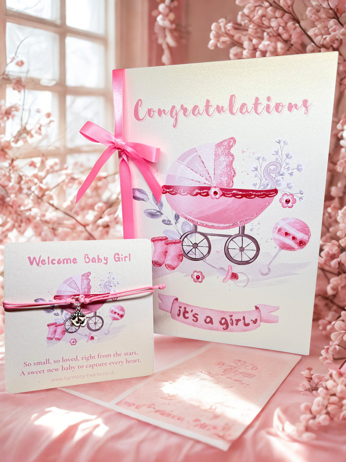 Pink baby shower card with stroller illustration and 'Congratulations it's a girl' text, with cord charm bracelet surrounded by pink flowers.